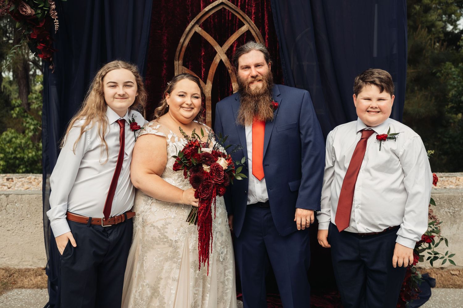 Newlyweds pose with two children at gothic arch altar after wedding ceremony.