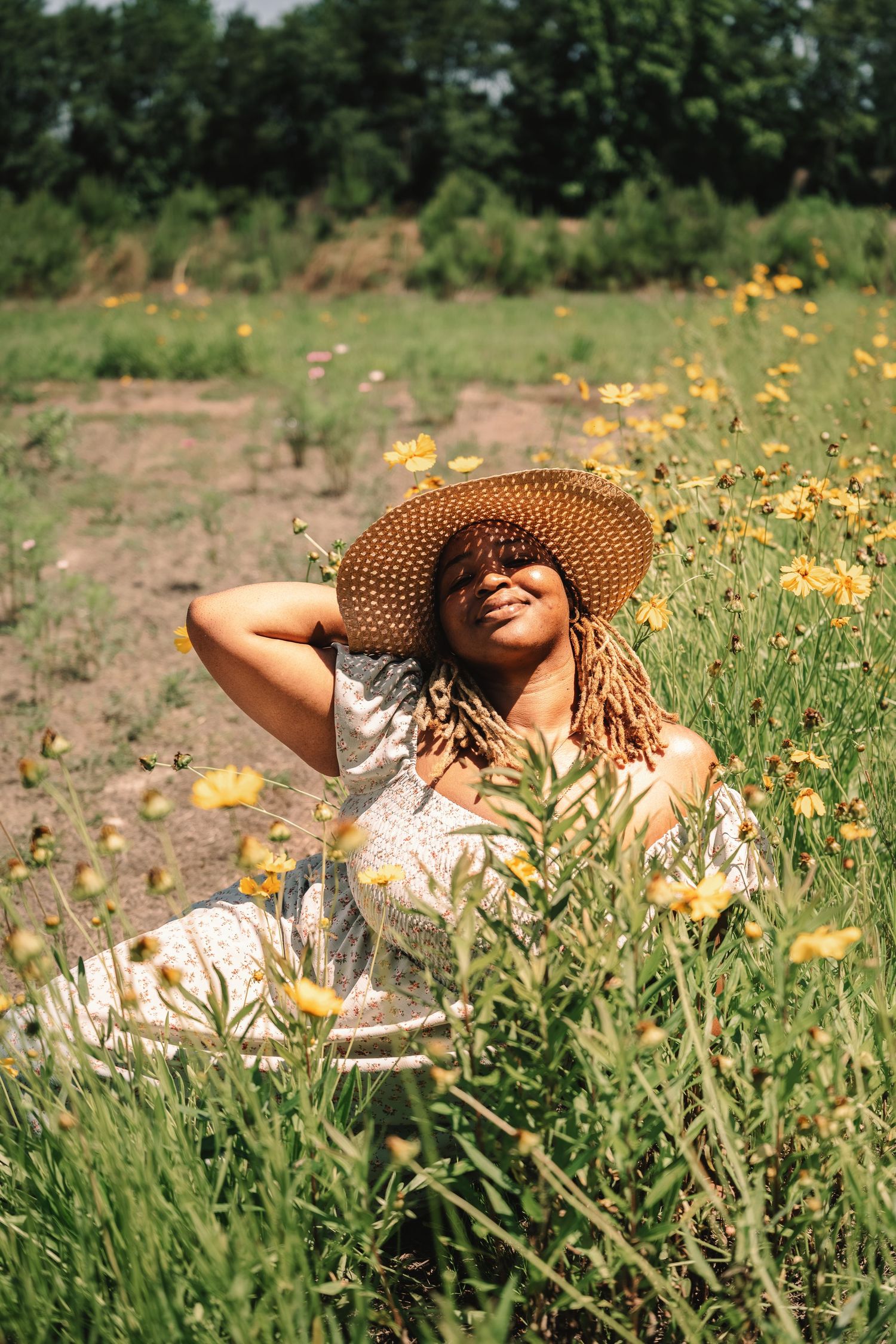 Woman in a floral dress and wide-brim straw hat relaxing in a wildflower meadow on a sunny day.