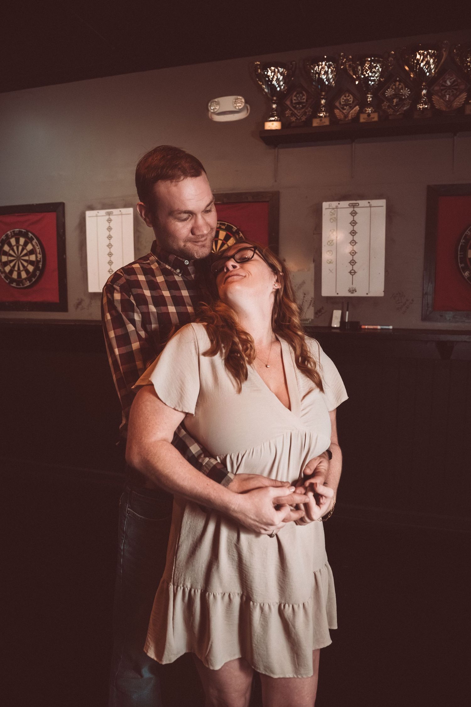A couple embracing tenderly in a bar setting with dartboards and trophies visible in the background.
