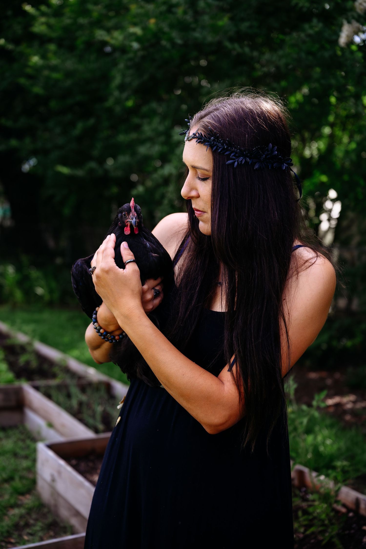 Young woman in black dress holds a black chicken in a lush green garden setting.