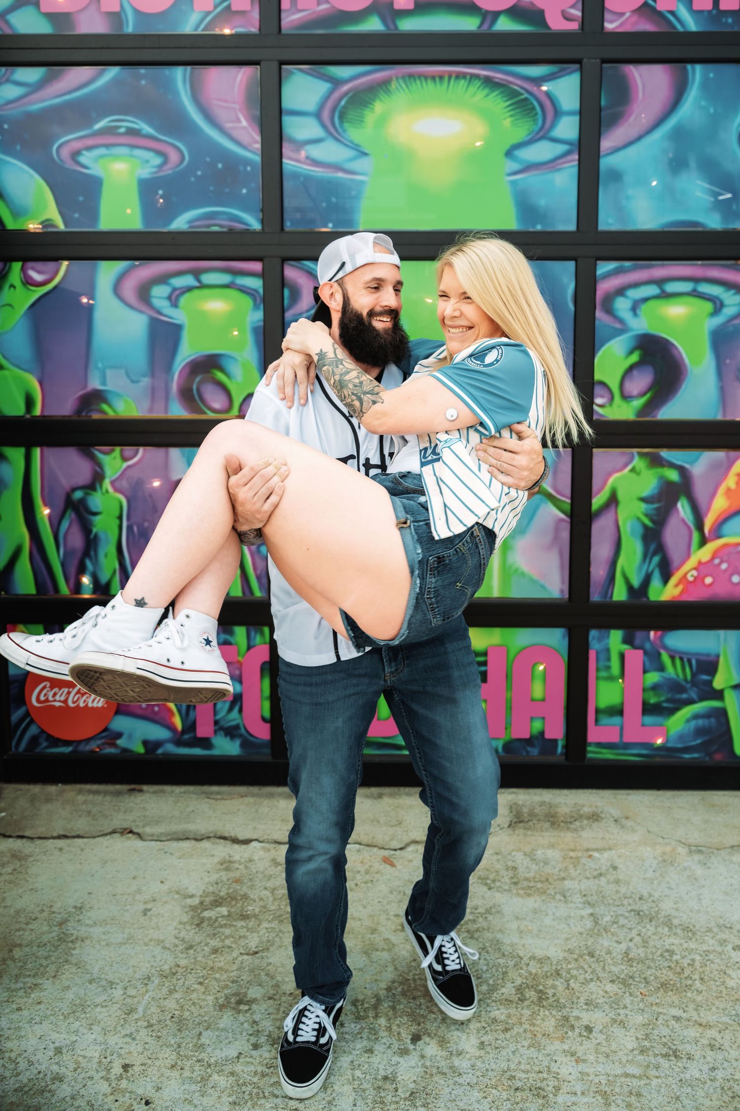 Tattooed bearded man carrying smiling blonde woman in his arms before colorful alien graffiti wall.