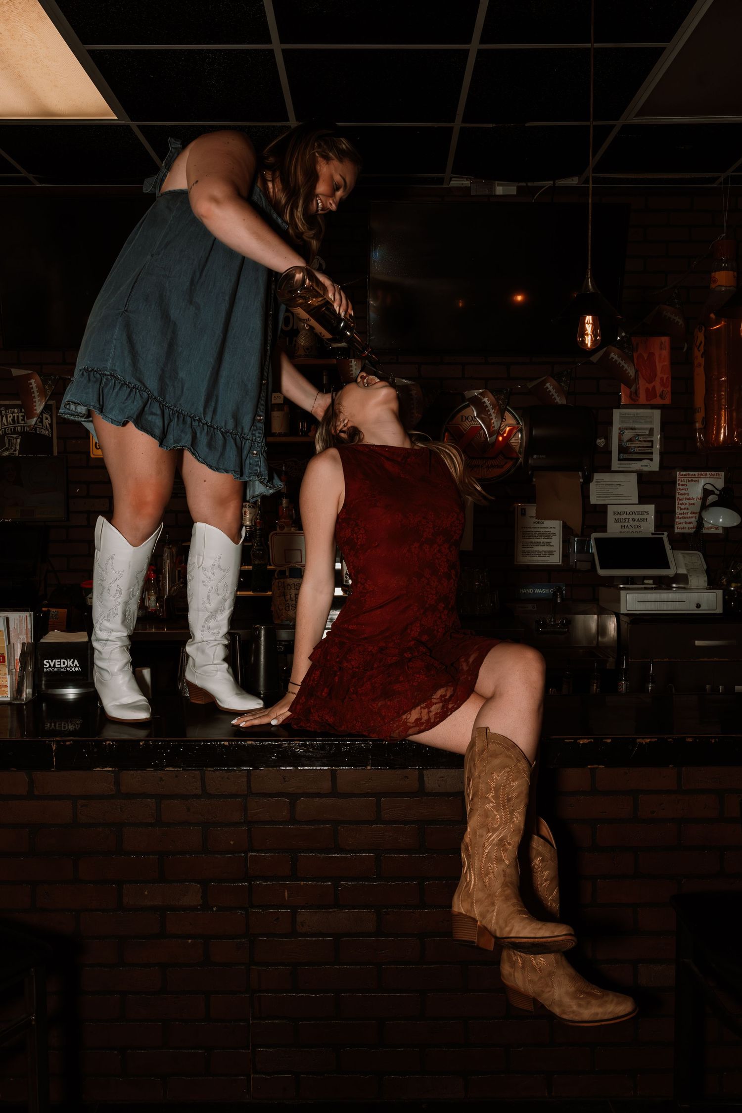 Two women posing dramatically on a brick bar counter in a dimly lit tavern.