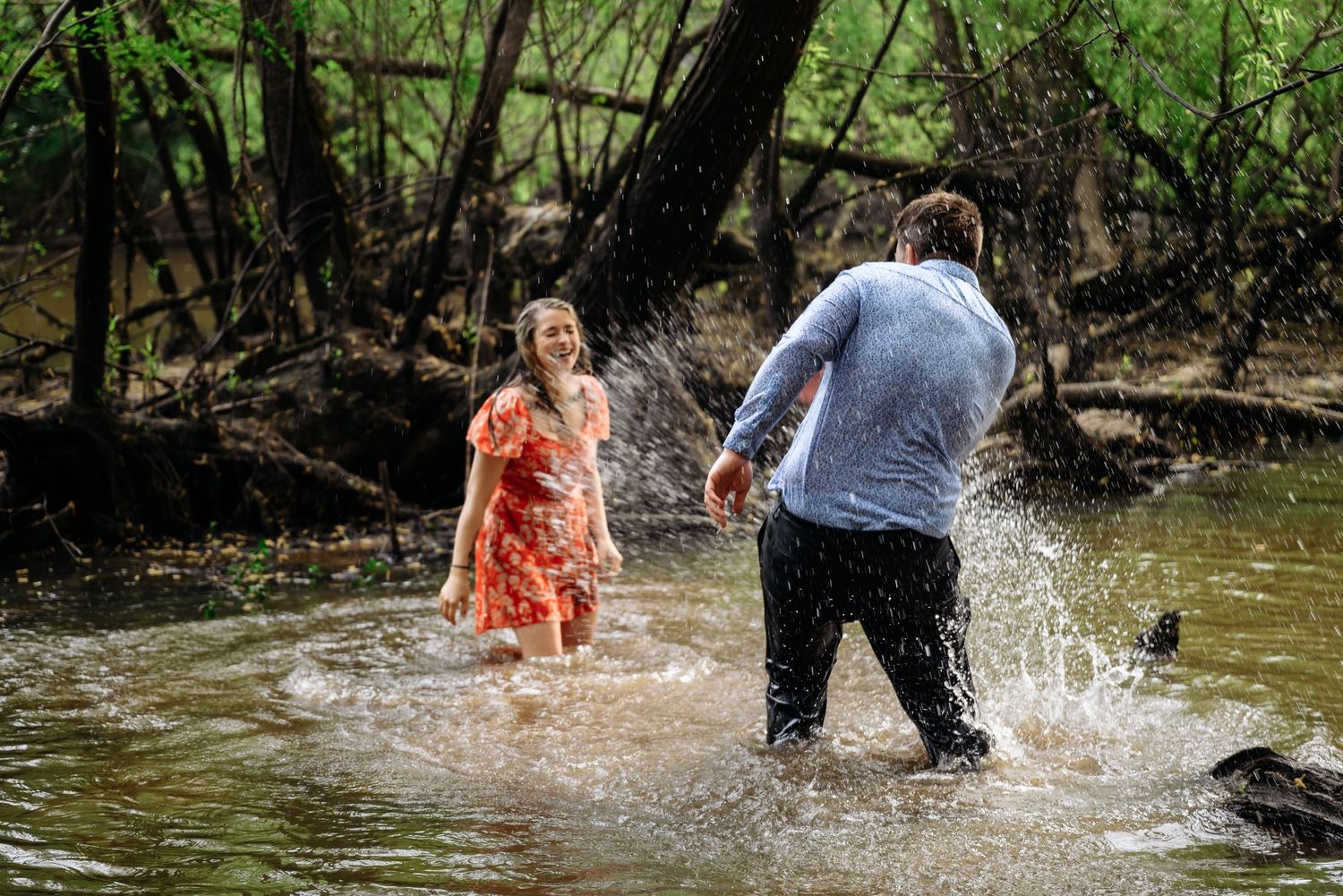 Couple splashing in a shallow creek surrounded by trees, woman in red floral dress laughing joyfully.