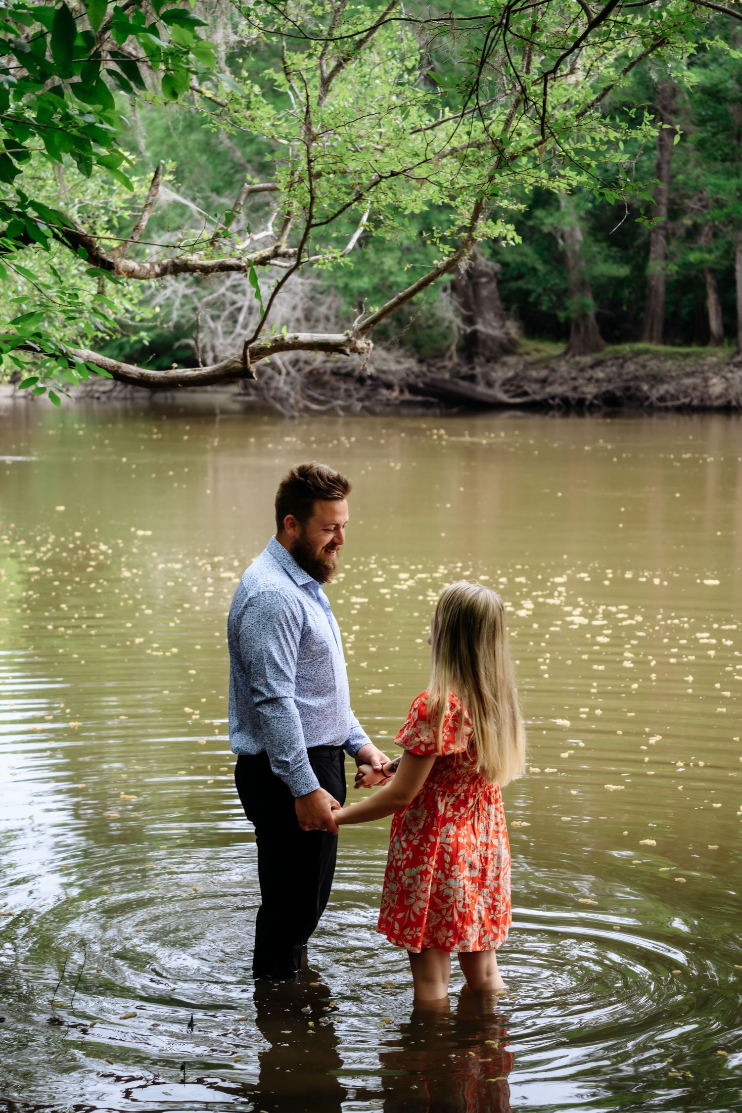 Bearded man in blue shirt holds hands with blonde woman in red floral dress while standing in a river.