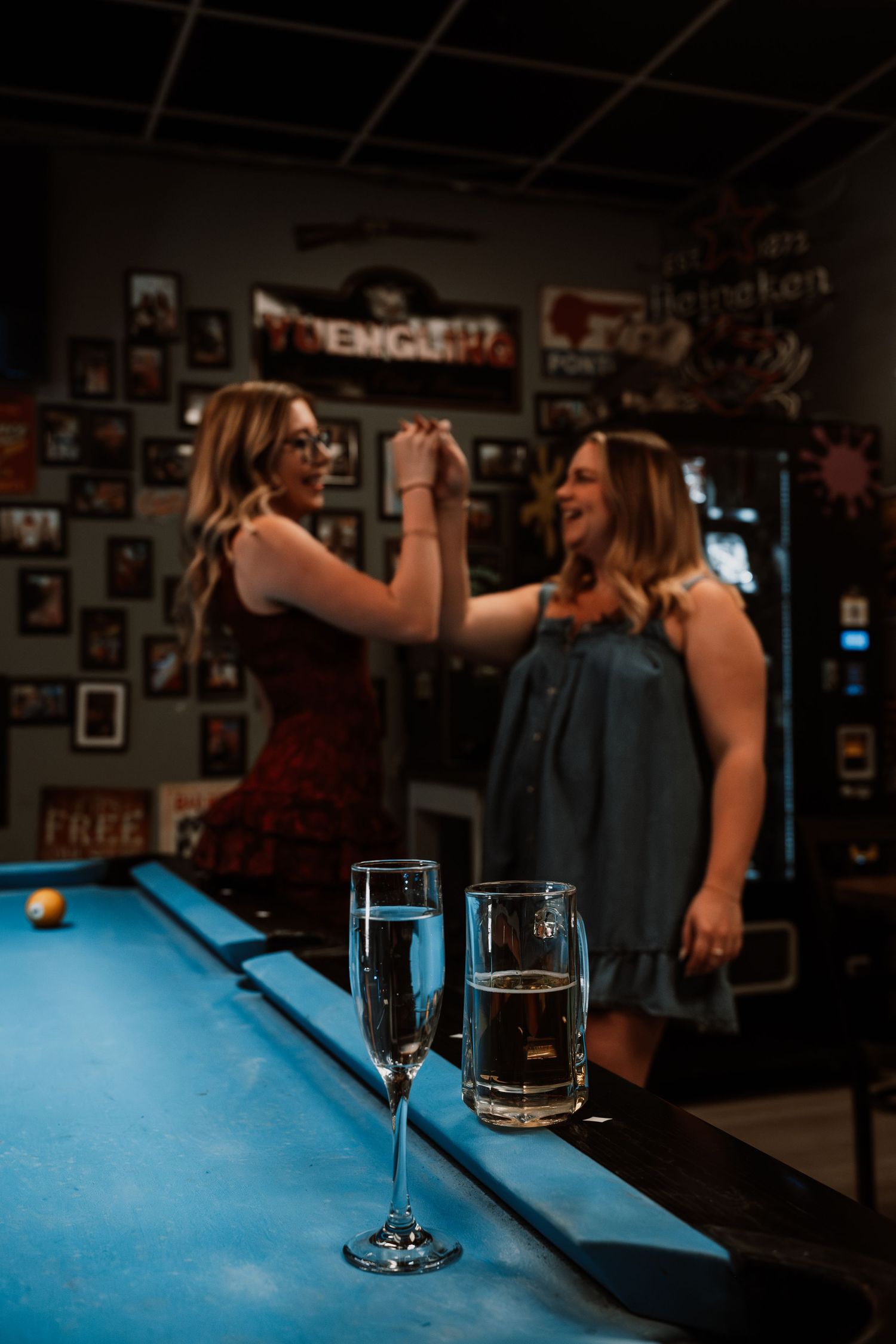 Two women dancing and celebrating near a pool table with drinks in a dimly lit bar.