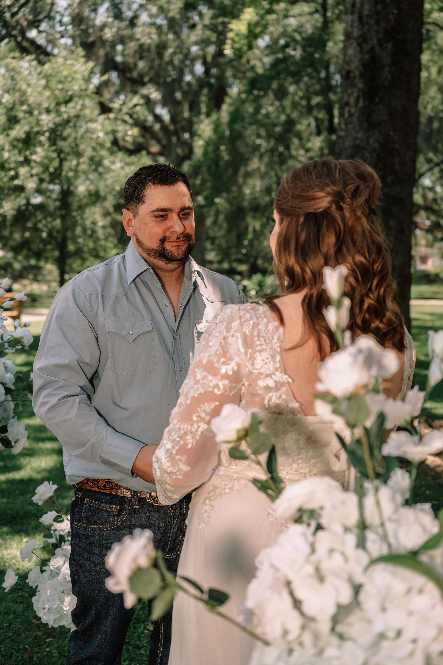 Bride and groom share a tender moment at outdoor wedding ceremony surrounded by white floral arrangements and lush green trees.