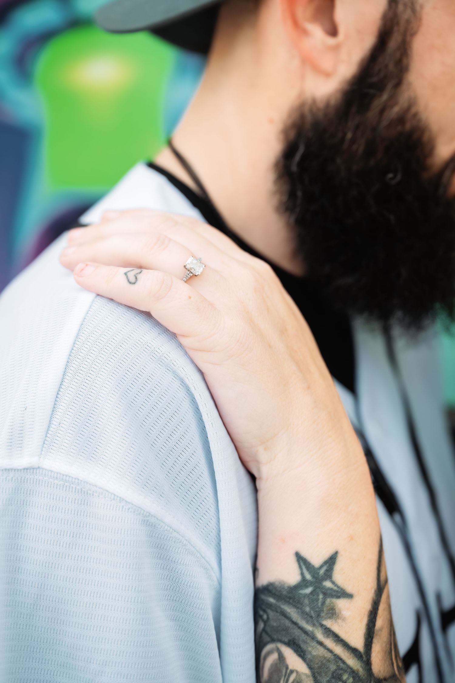 Close-up of woman's hand with engagement ring resting on bearded tattooed man's shoulder.
