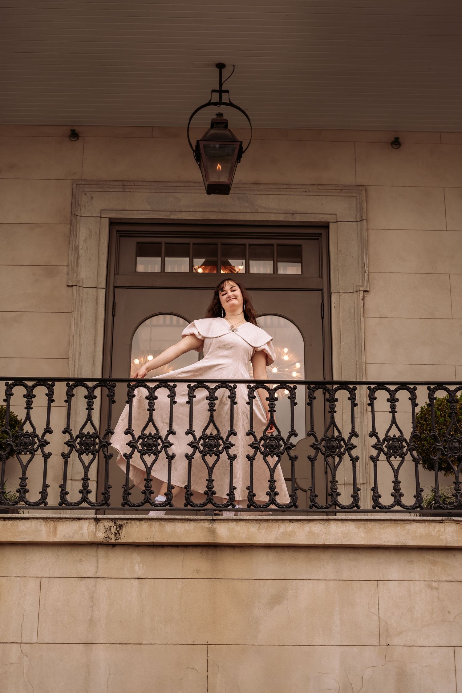 Woman in elegant white dress with cape posing on an ornate iron balcony of a building.