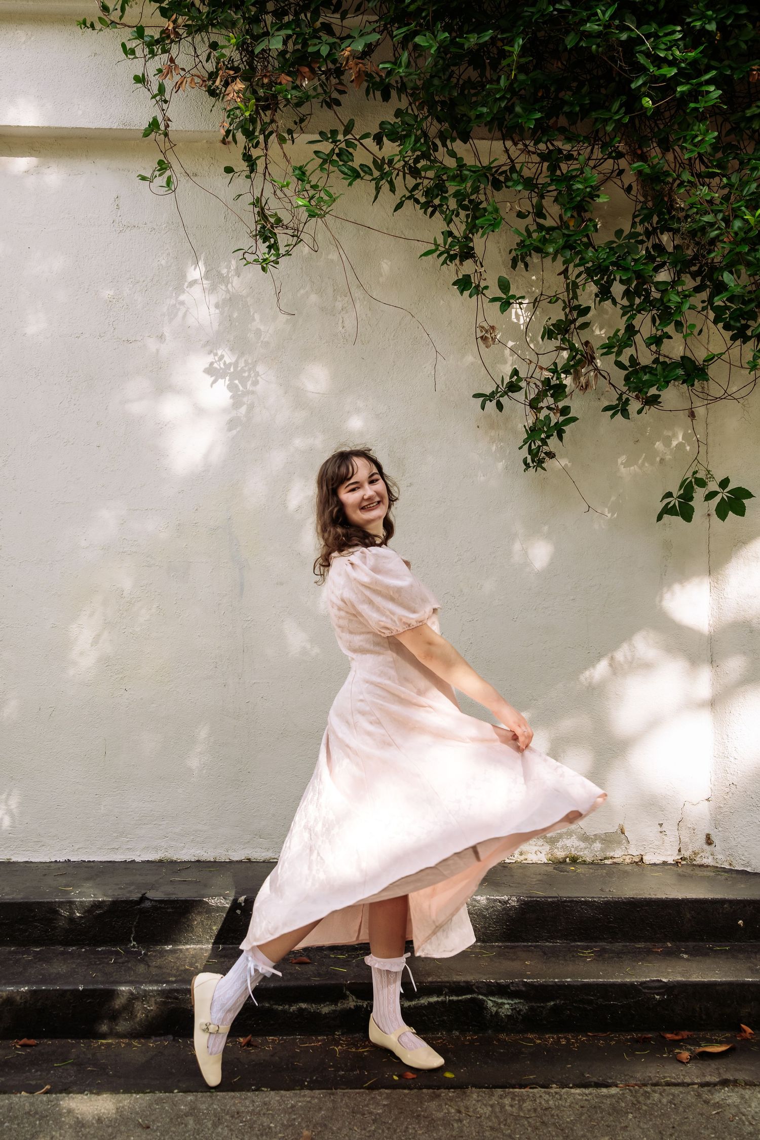 Smiling woman in light pink midi dress twirling on steps beside a white wall with ivy.