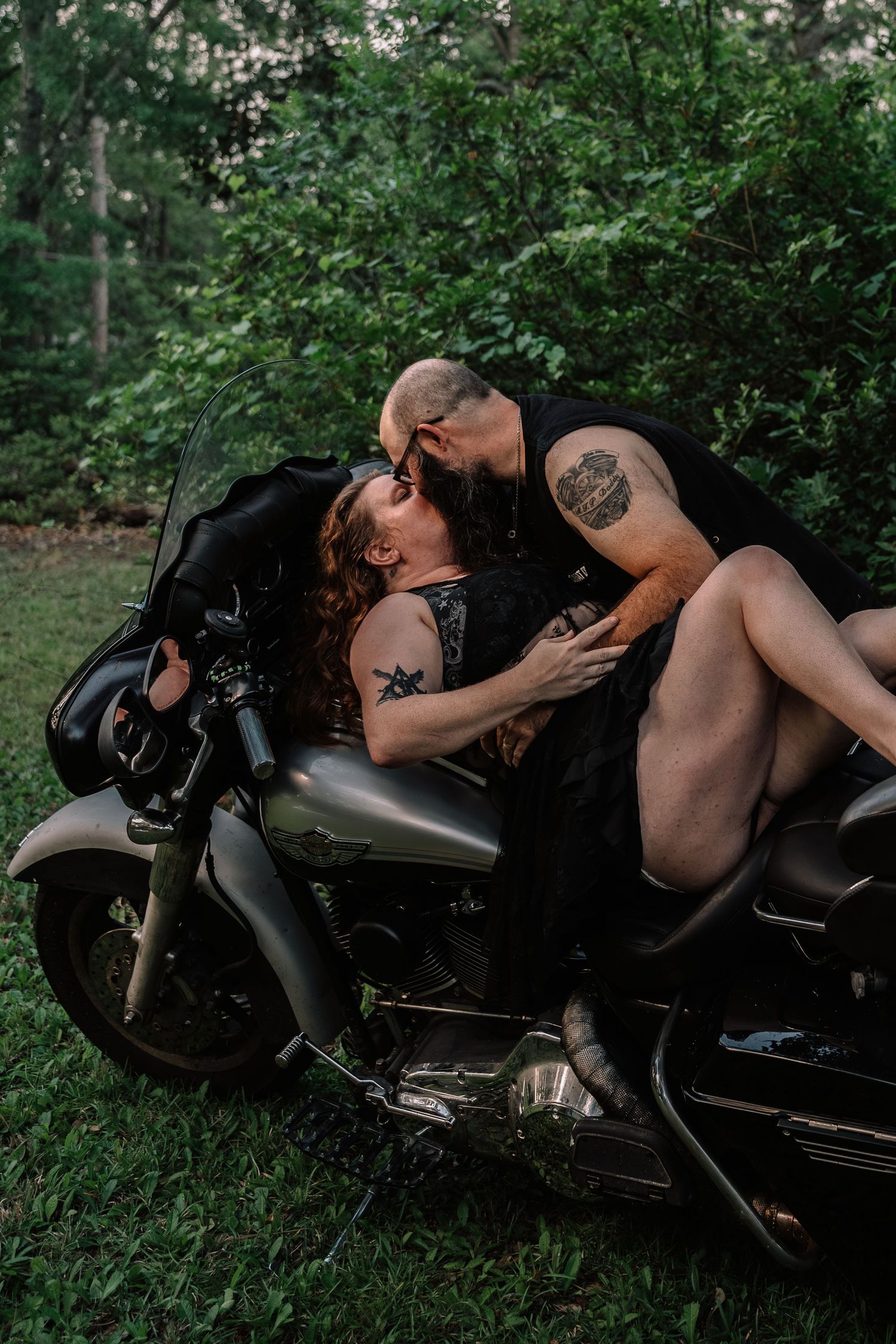 Tattooed couple kissing passionately on a black motorcycle surrounded by green foliage.