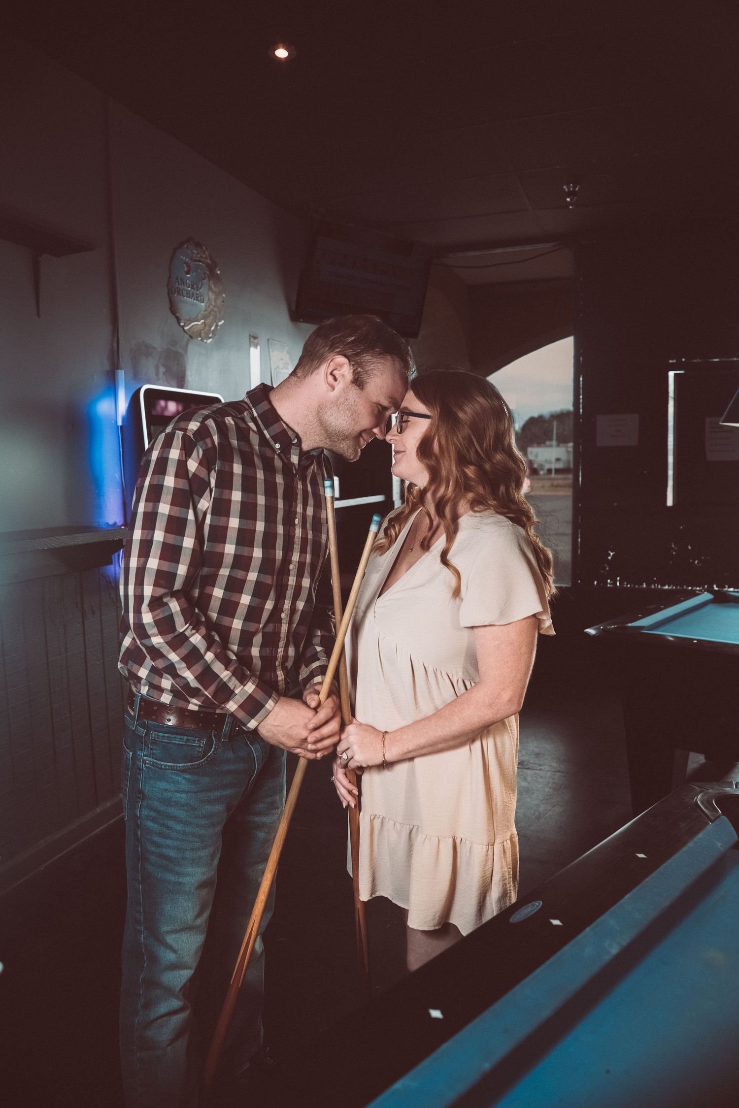 A romantic couple shares an intimate moment while playing pool in a dimly lit game room.