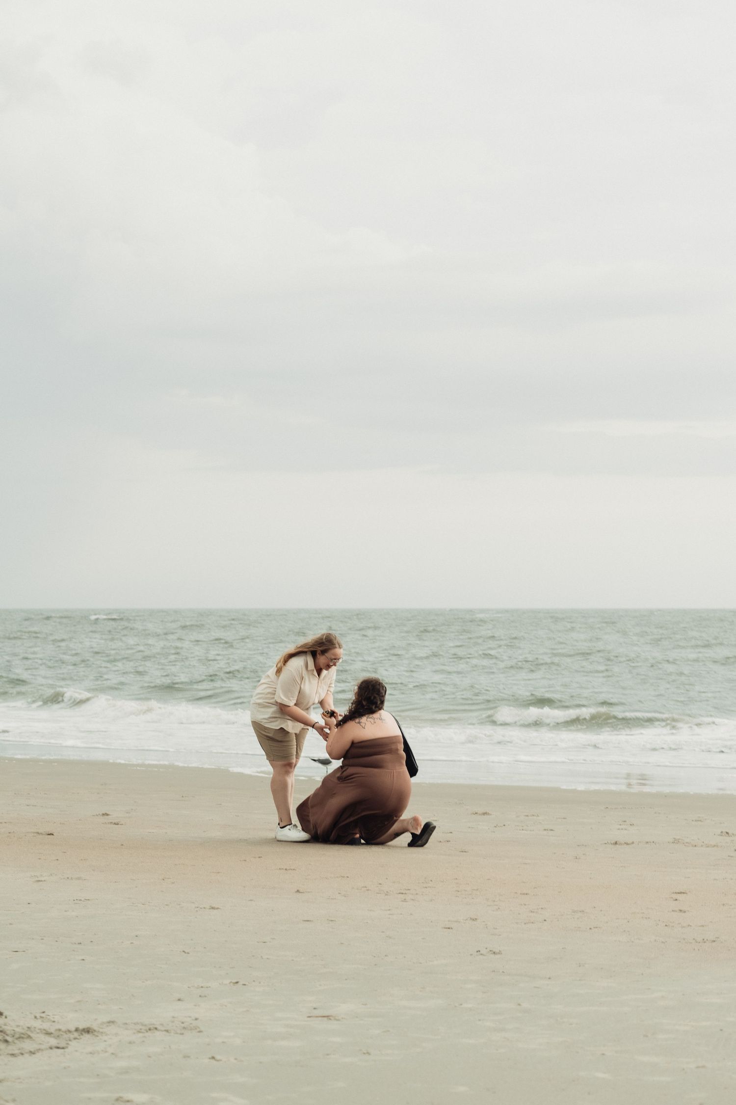 Two women on a sandy beach, one sitting and one leaning down to help the other up near the ocean.