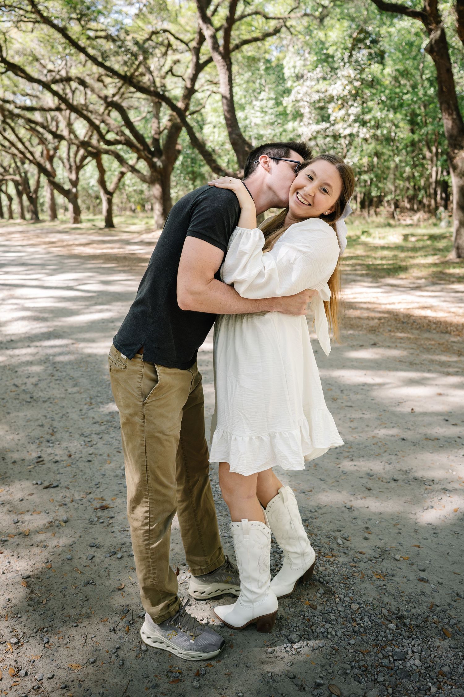 Man kissing womans cheek as she laughs joyfully while wearing white dress and cowboy boots.