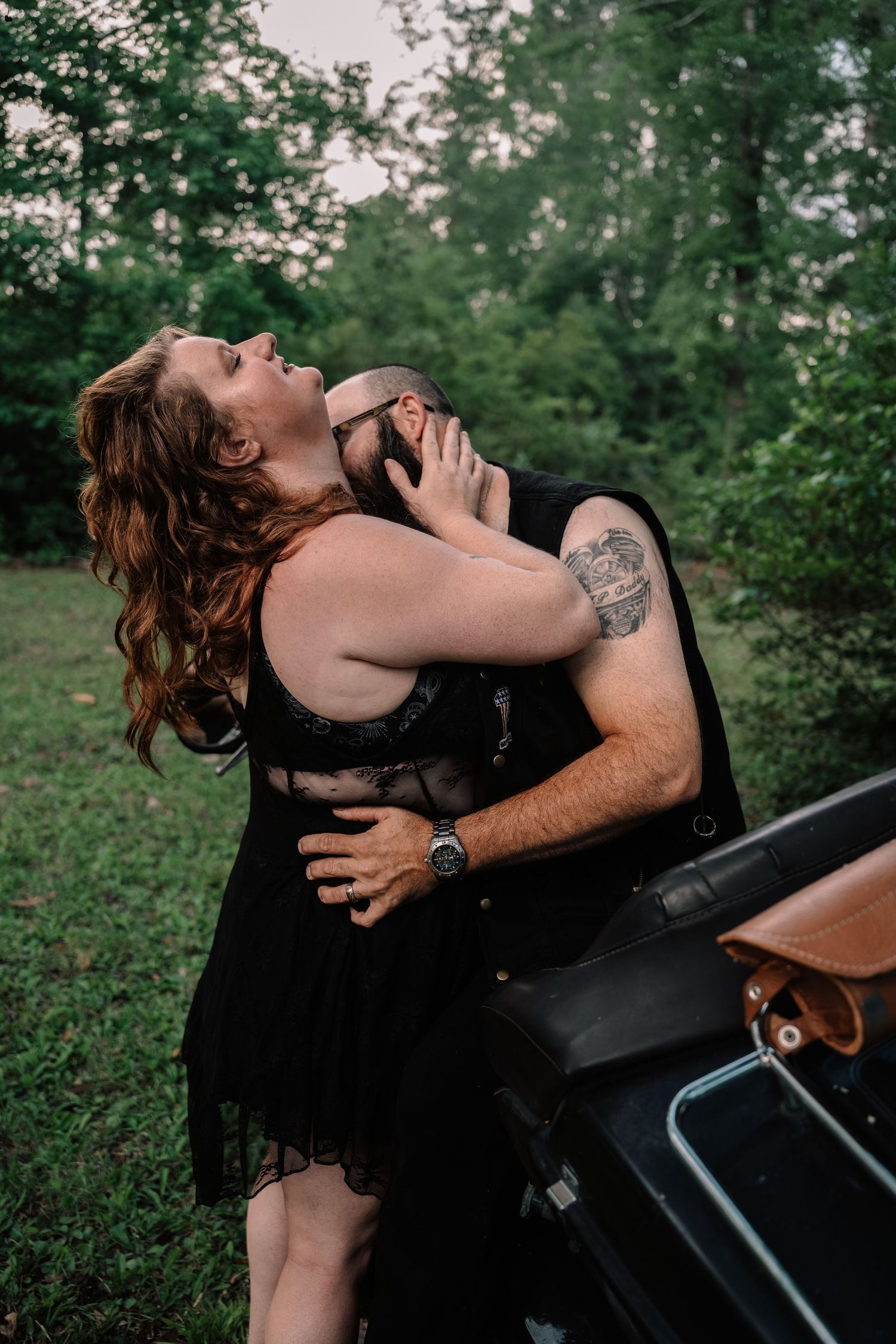Couple embracing passionately near a motorcycle in a lush green forest setting.
