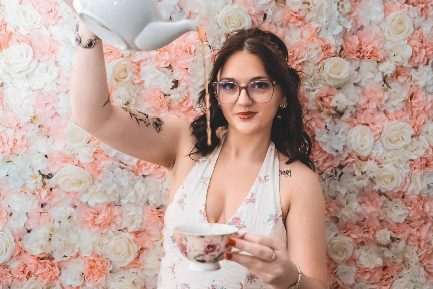 A person in glasses and white dress pours tea against a pink floral backdrop.