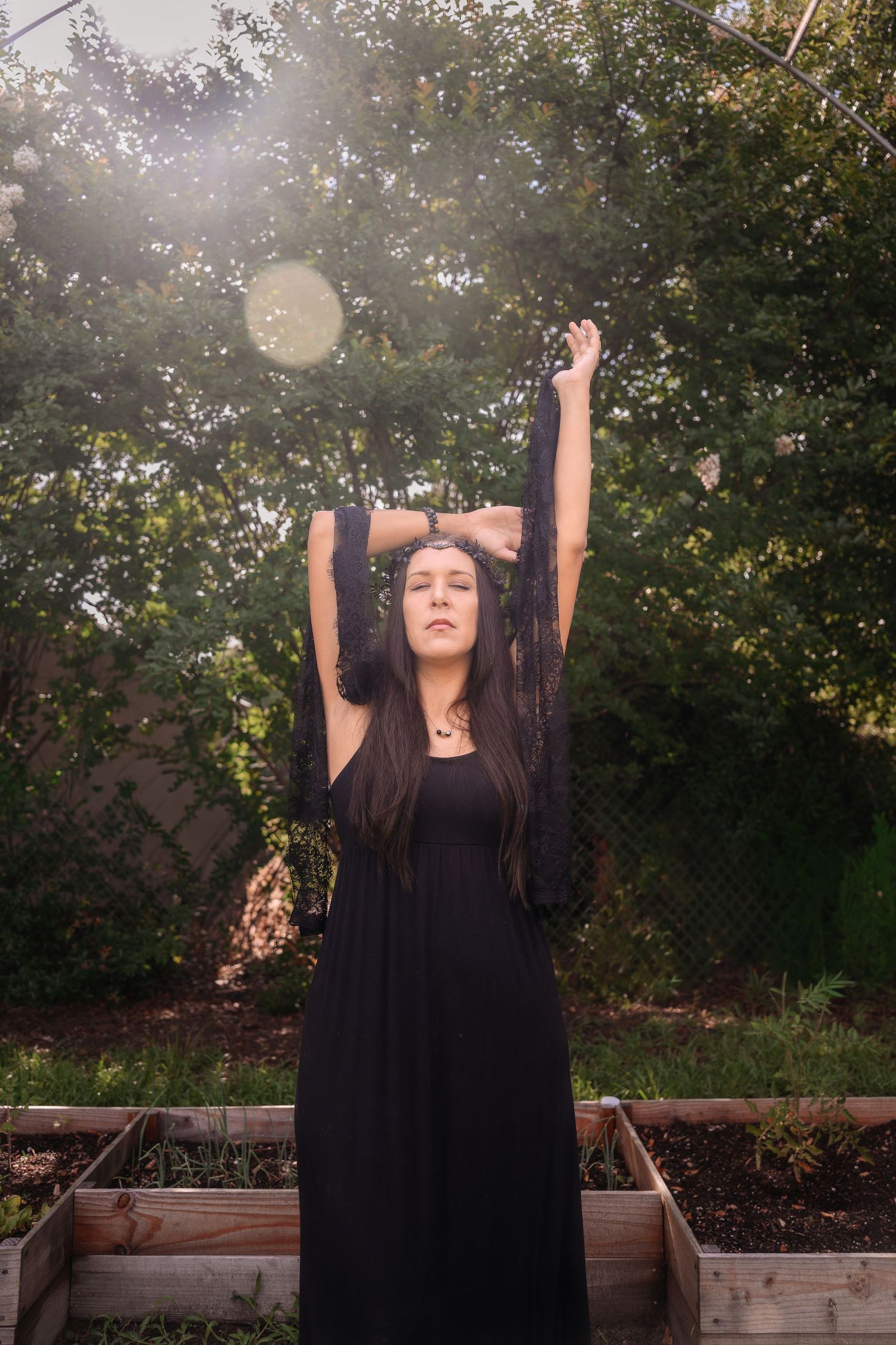 Woman in black dress raises arms in sunlit backyard garden with raised planting beds.