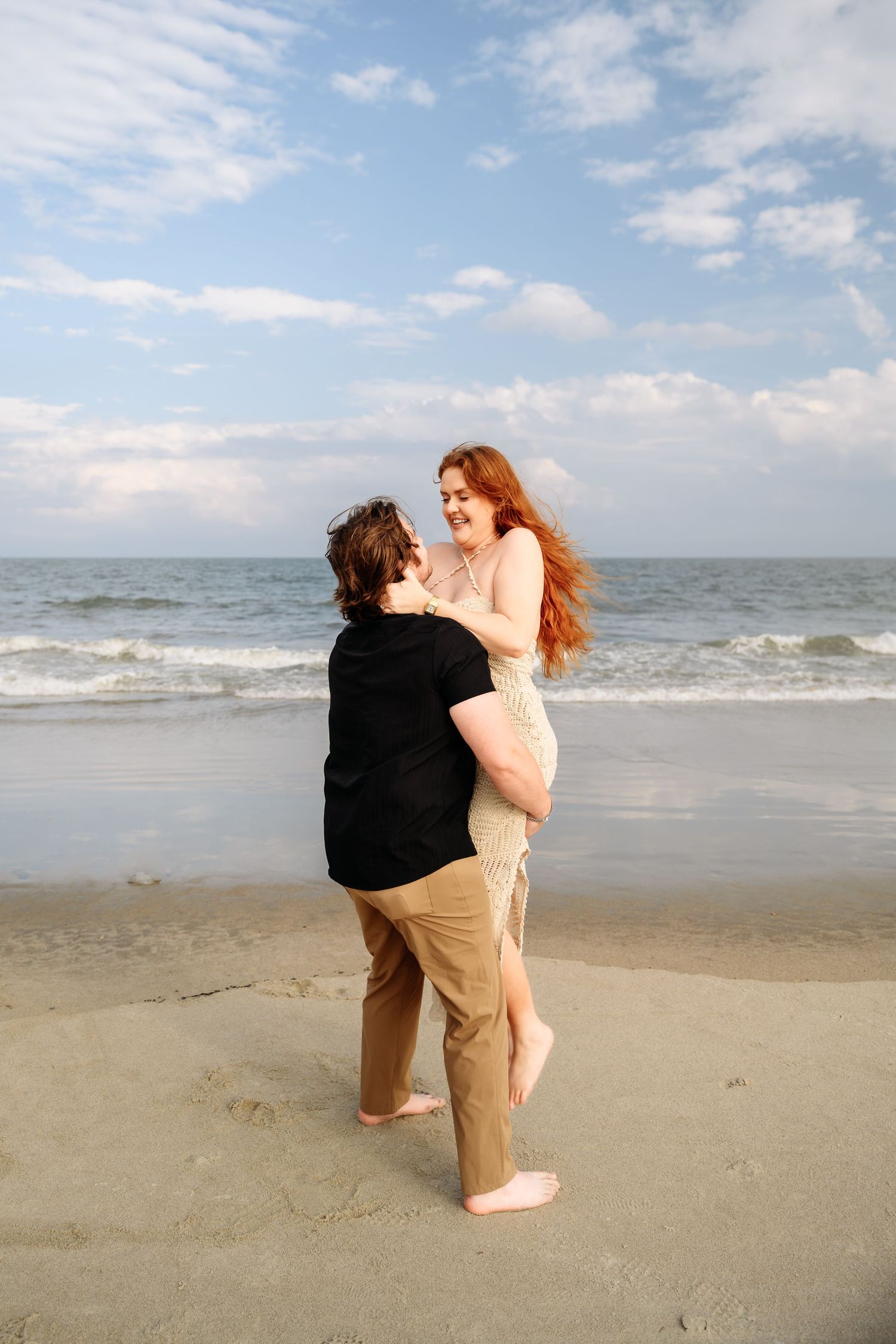 Man lifting and spinning redhead woman joyfully on a sandy beach after beach proposal.
