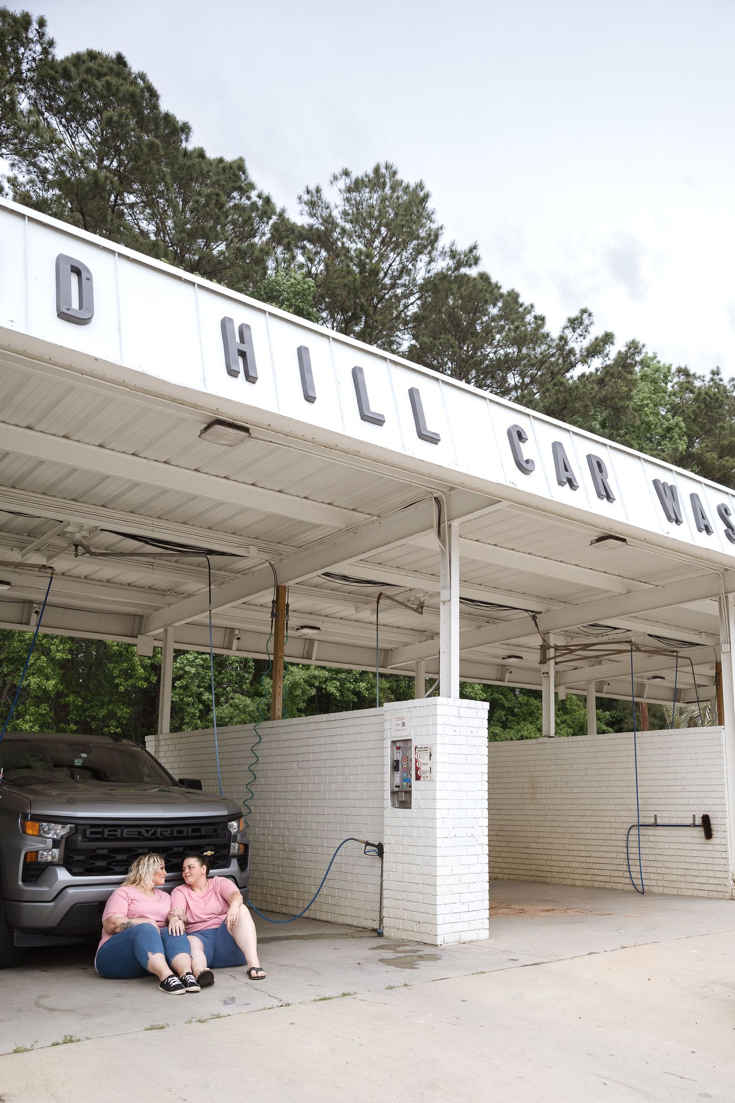 Two women in matching pink shirts sitting under a car wash canopy by a truck.