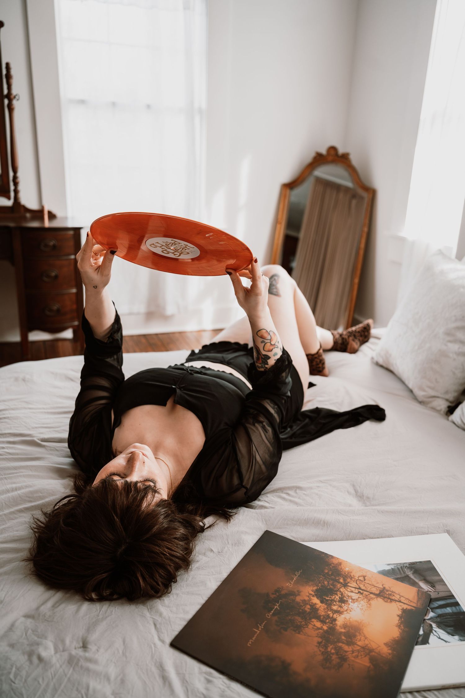 Woman in black outfit lies on bed holding orange vinyl record up in sunlit room.