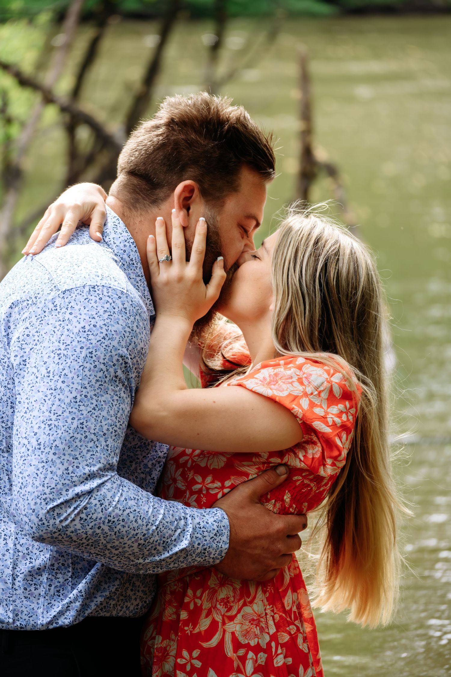 Couple sharing a romantic kiss outdoors near a river, woman in red floral dress with engagement ring visible.