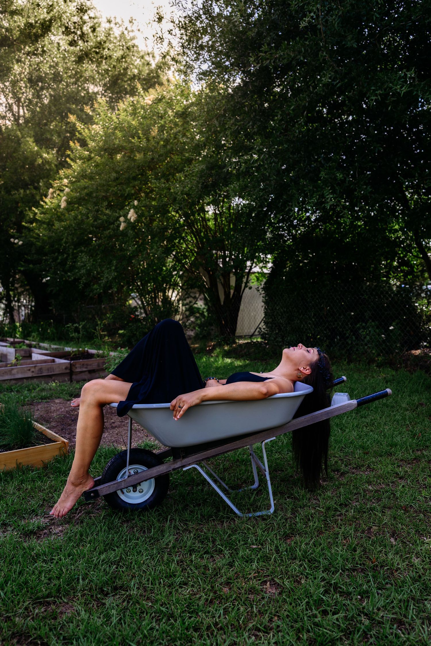 Woman in black dress relaxing in a wheelbarrow in a green backyard garden setting.