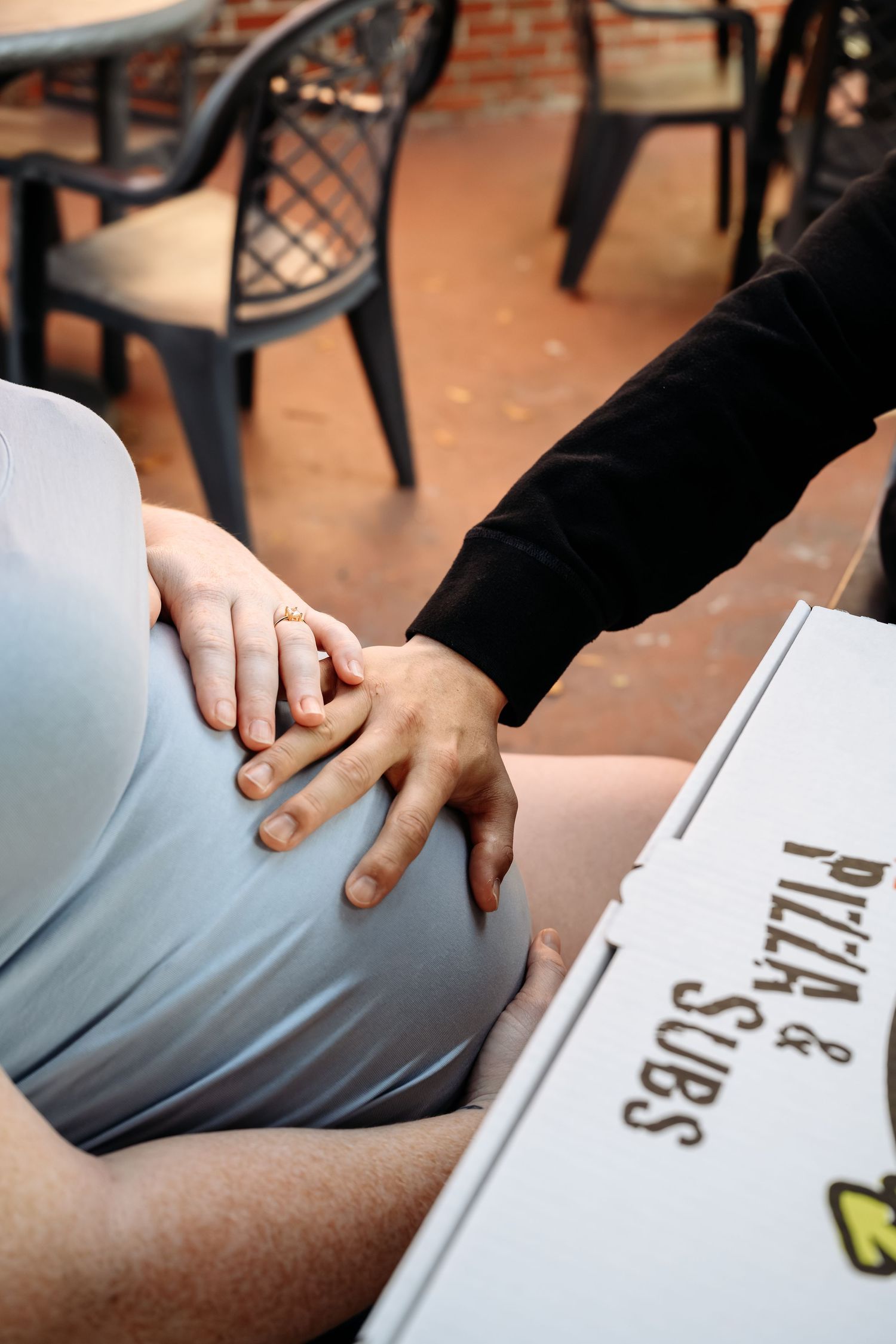 Close-up of a couple holding hands over a pregnant belly beside a Pizza and Subs box at an outdoor table.