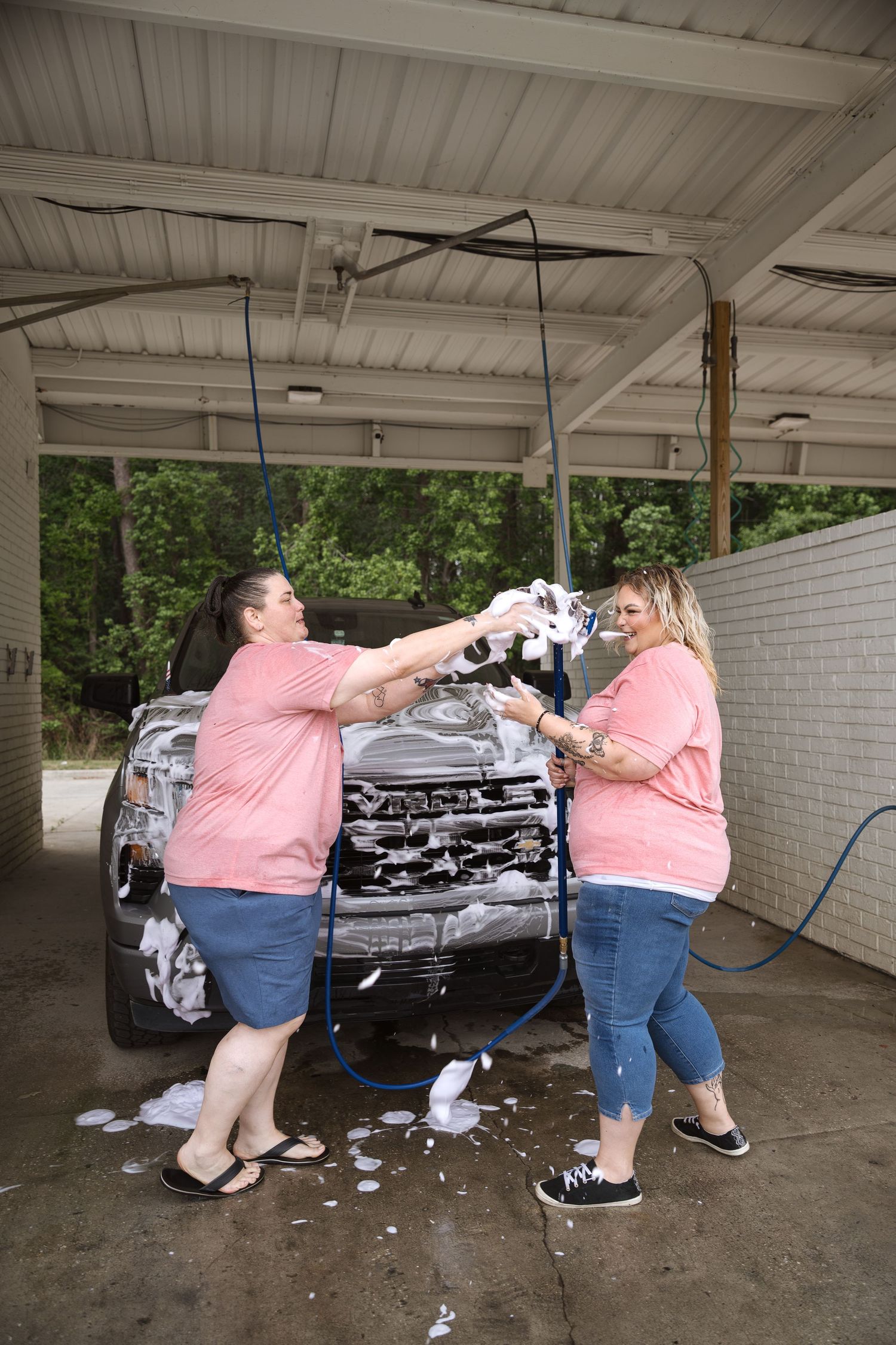 Two women in pink shirts playfully throwing soapy sponges while washing a truck.
