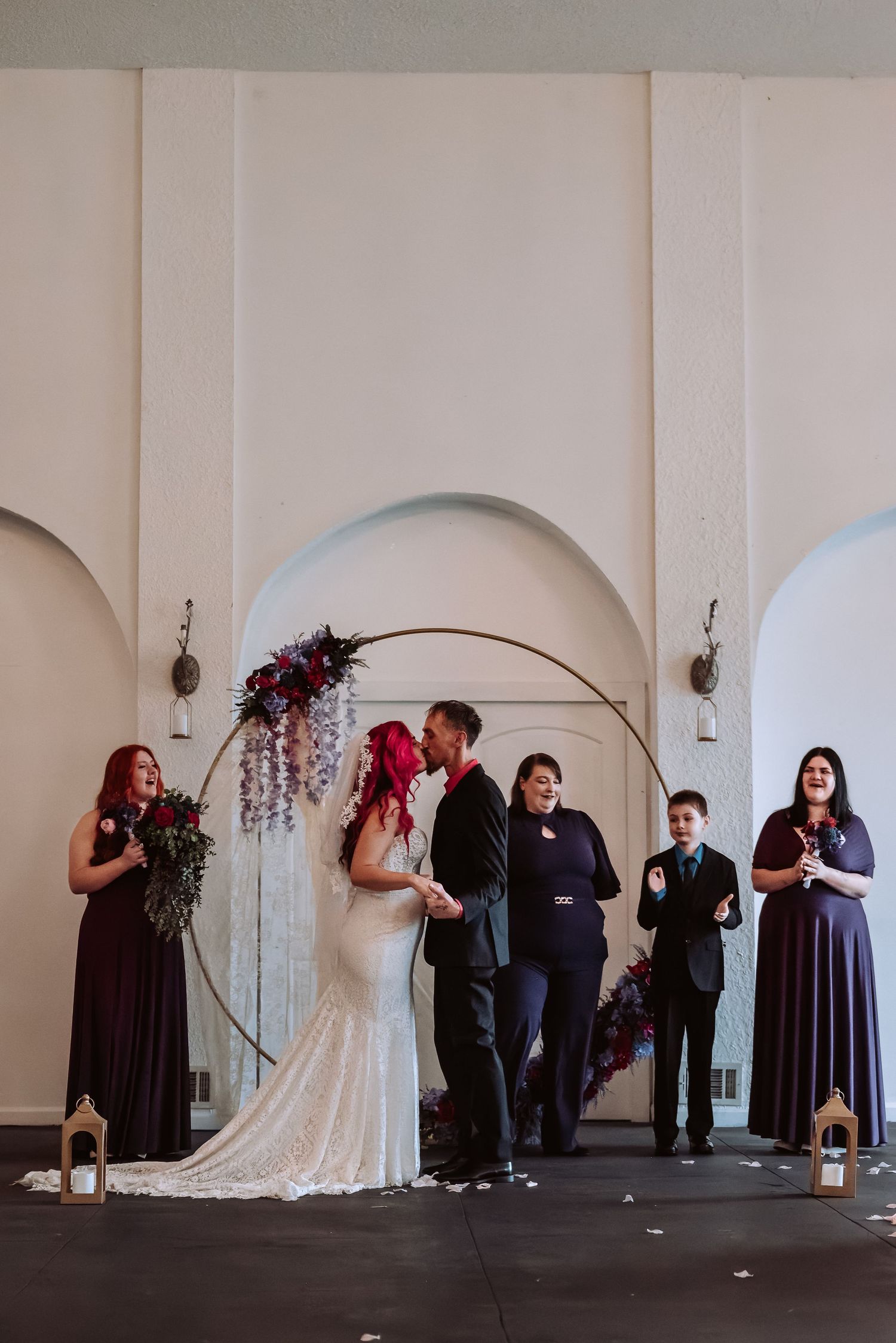 Bride and groom share first kiss under a floral arch with wedding party in purple dresses at ceremony.