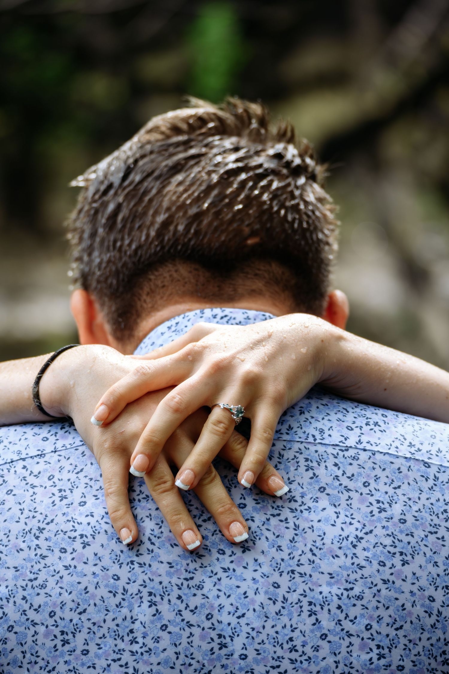 Close-up of a woman's hands with an engagement ring wrapped around a man's shoulders in a tender embrace.
