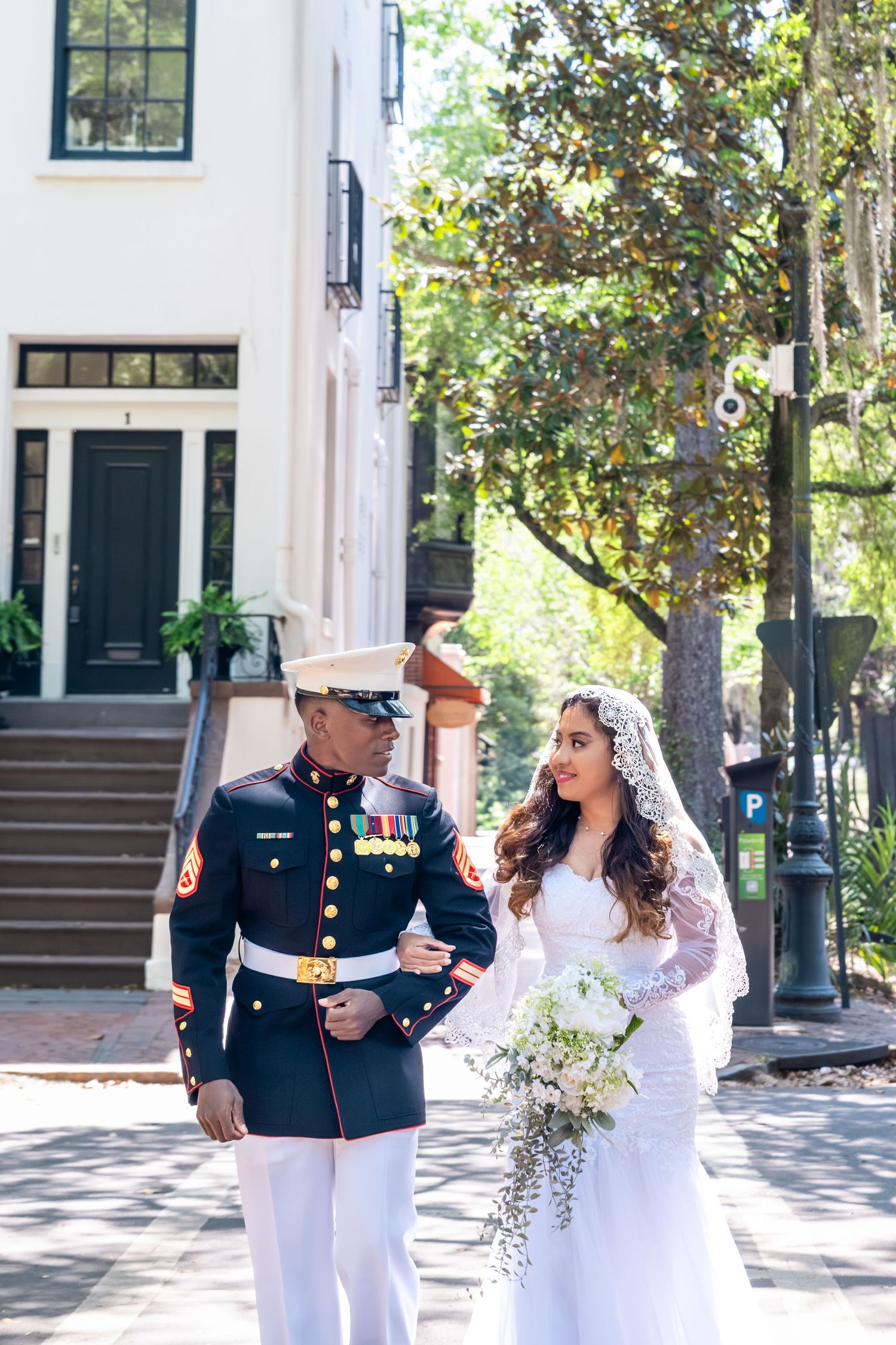 Marine groom in dress blues escorts beautiful bride in lace gown along a sunny tree-lined city street.