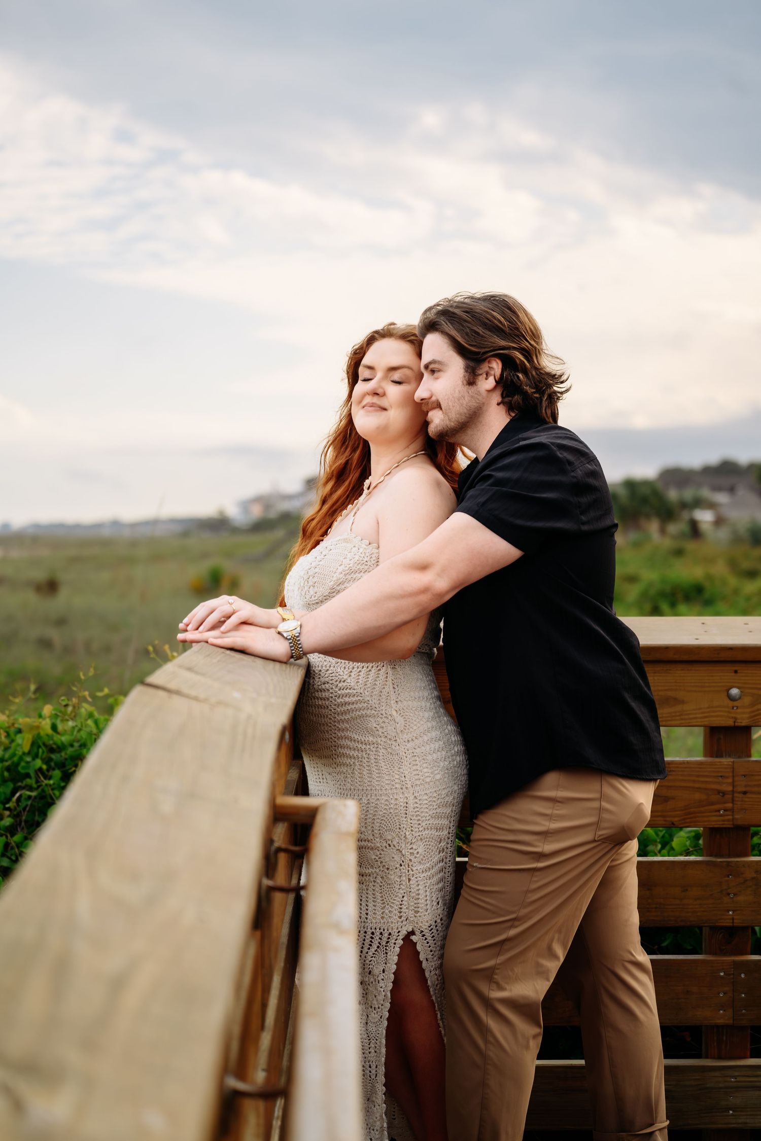 Romantic couple embracing on a wooden deck overlooking a green coastal landscape at sunset.