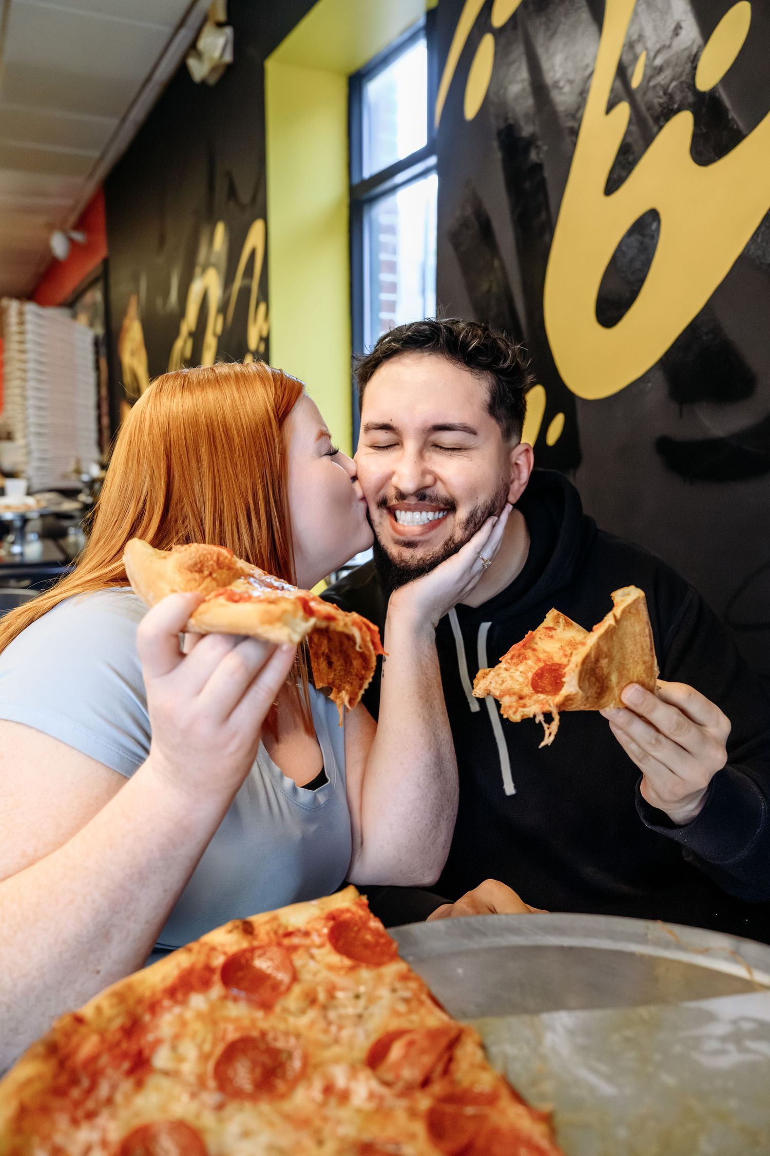 Smiling couple sharing a romantic moment while eating pepperoni pizza slices inside a pizzeria restaurant.