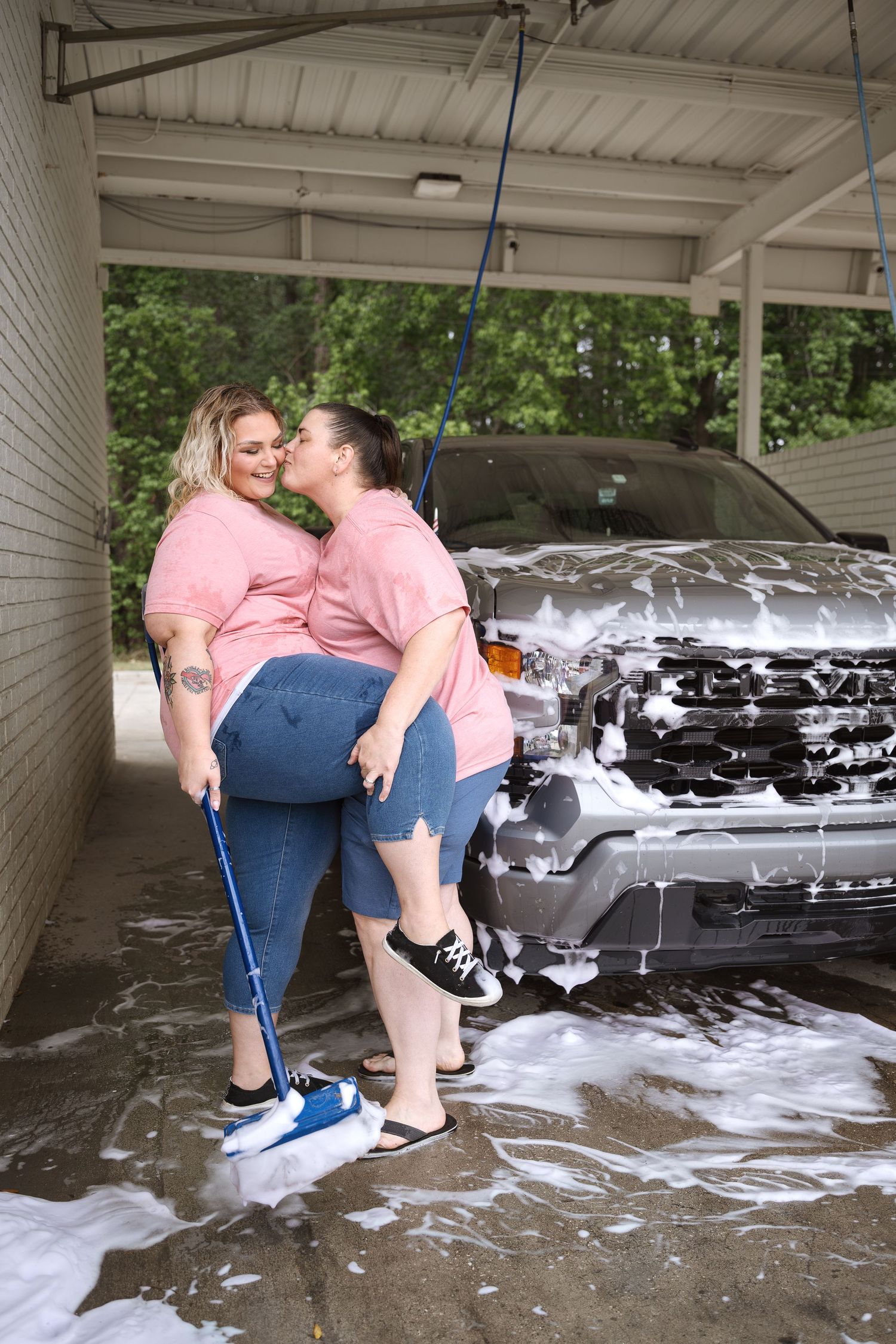 Two women in matching pink shirts share a kiss while washing a truck covered in soap suds at a self-service car wash.