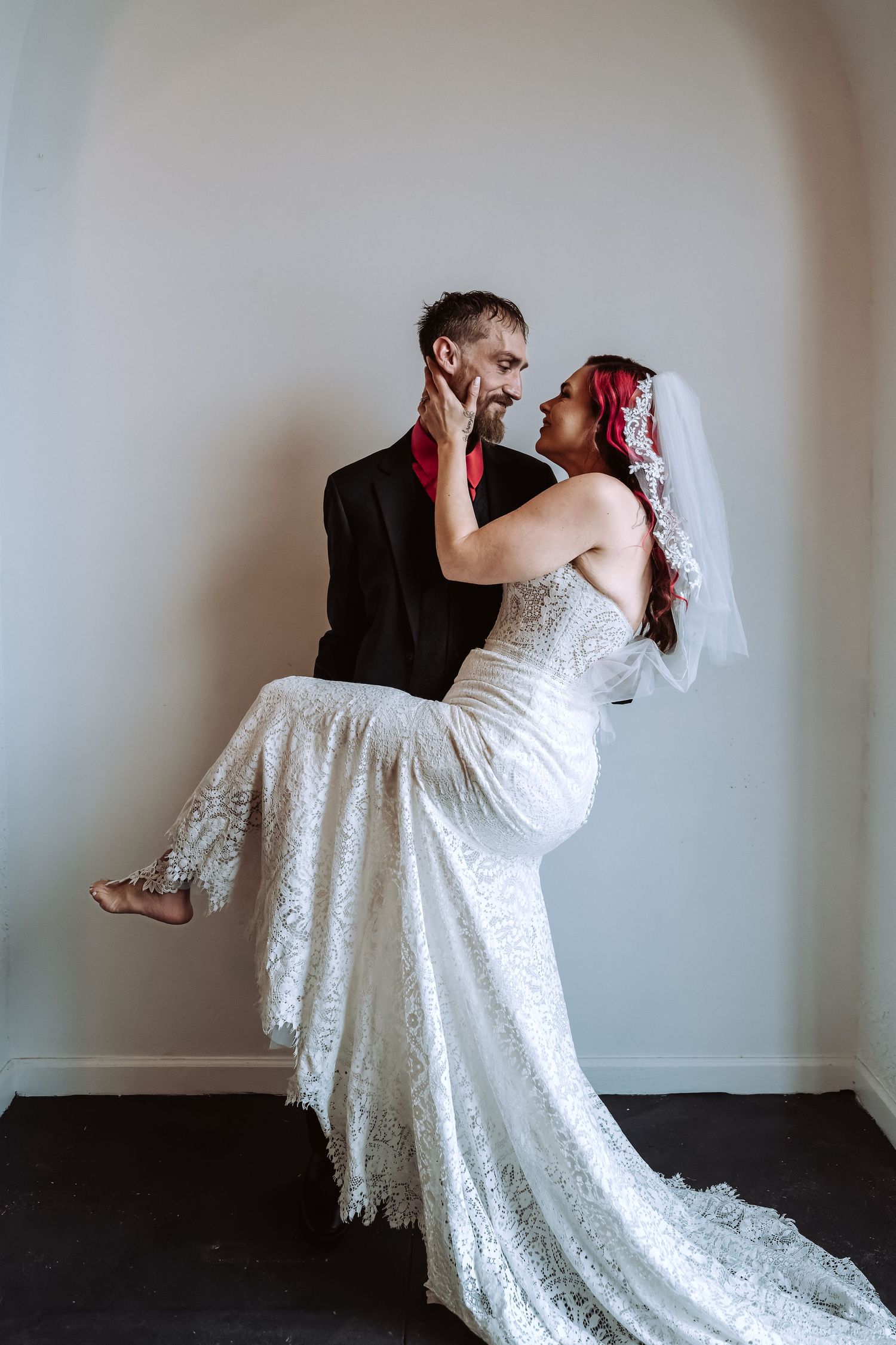 Groom lifting bride in a lace wedding dress as they share a romantic moment against a white wall.
