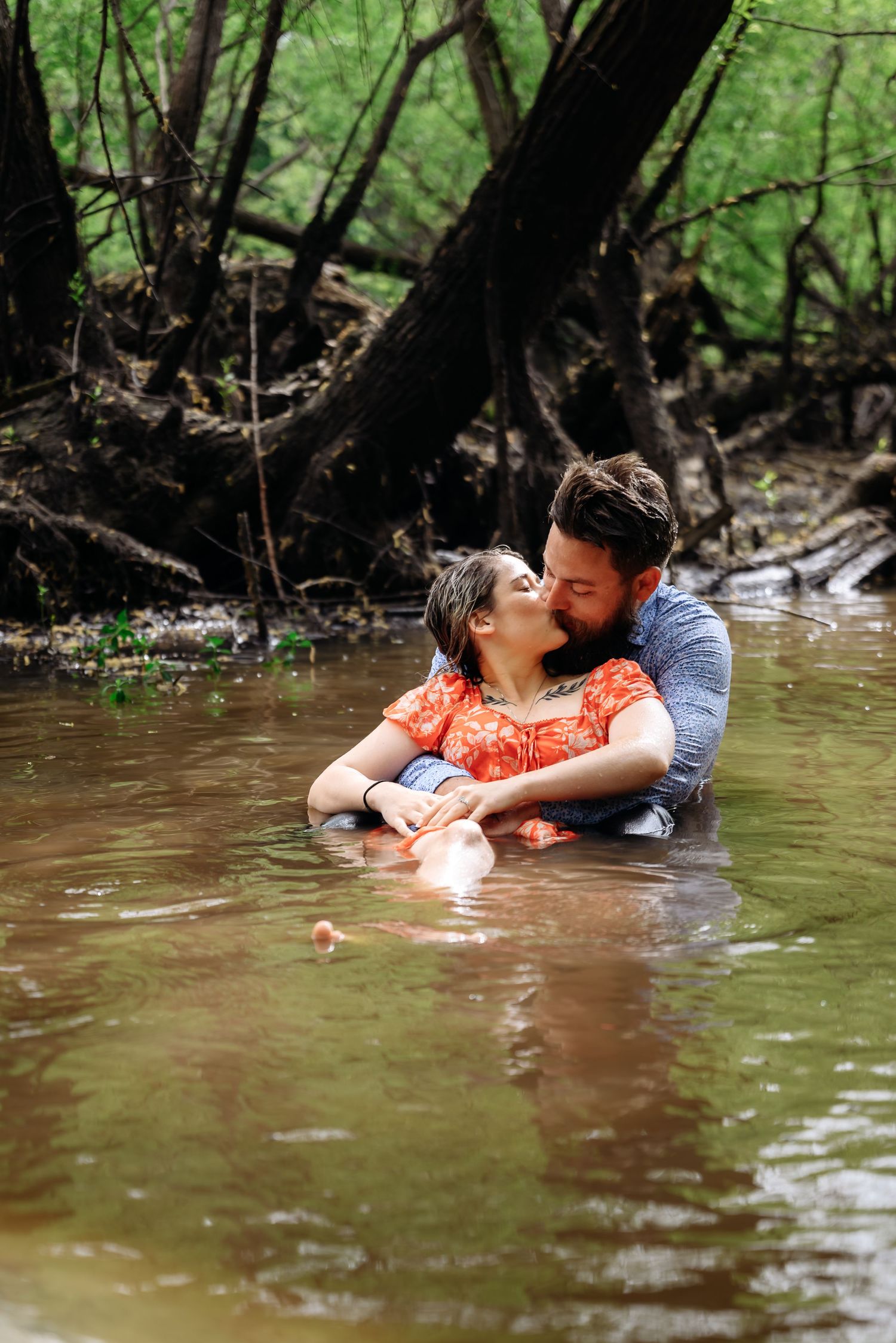 Couple embracing romantically while standing in a shallow forest creek surrounded by lush green trees.