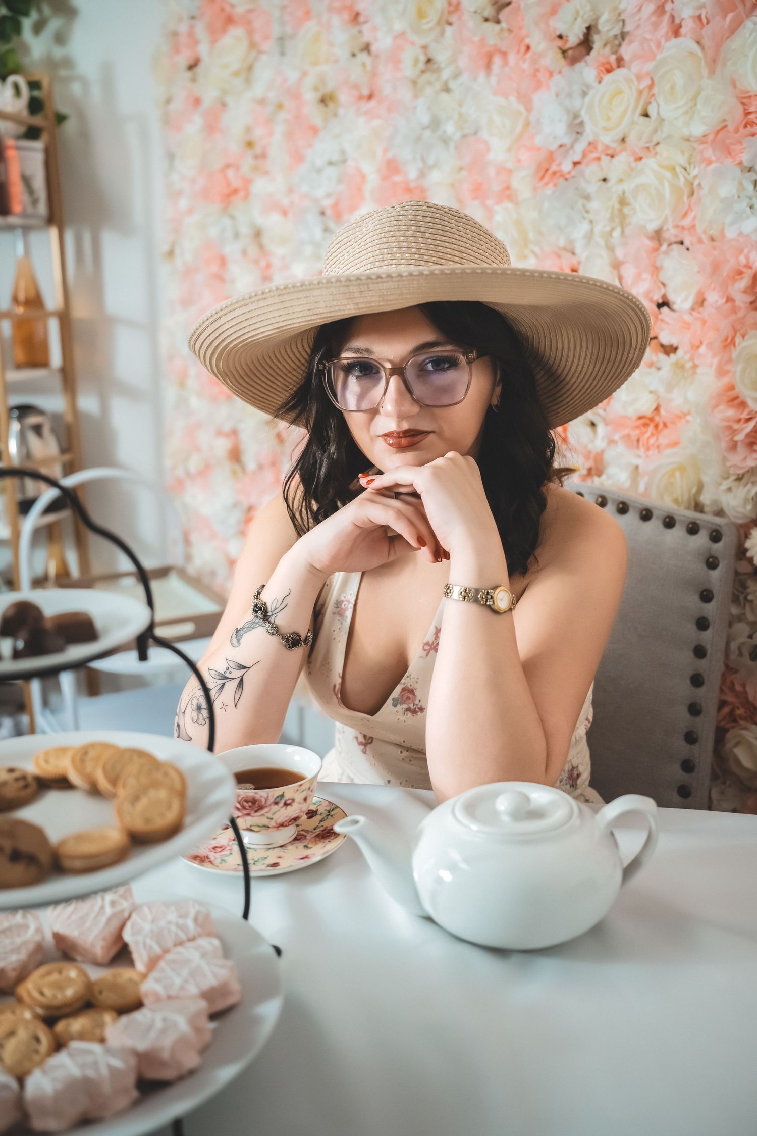 A tea party setting with floral backdrop, teapot, and pastries on a table with someone wearing a sun hat and glasses.