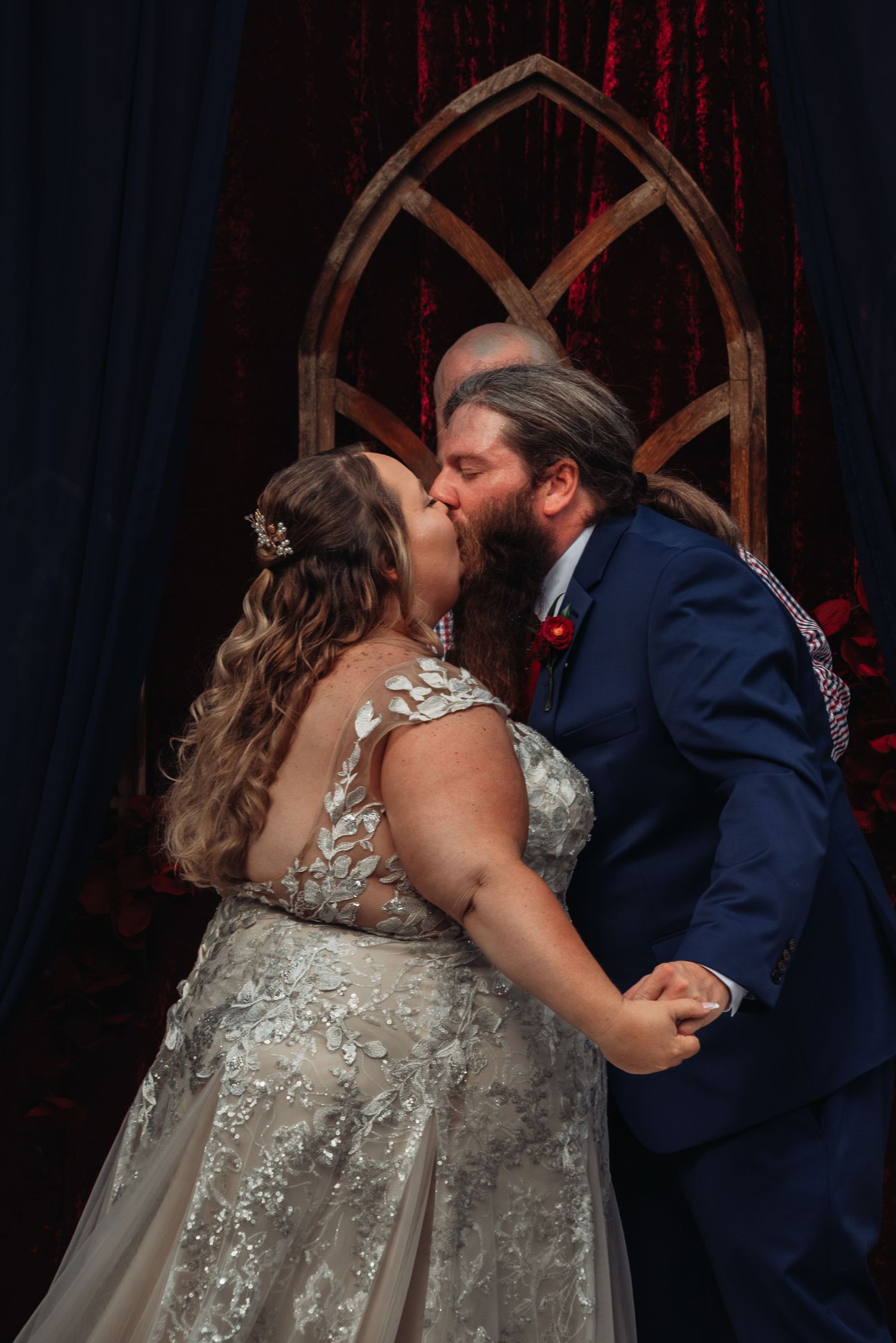 Bride and groom share first kiss at gothic arch altar with red velvet backdrop.