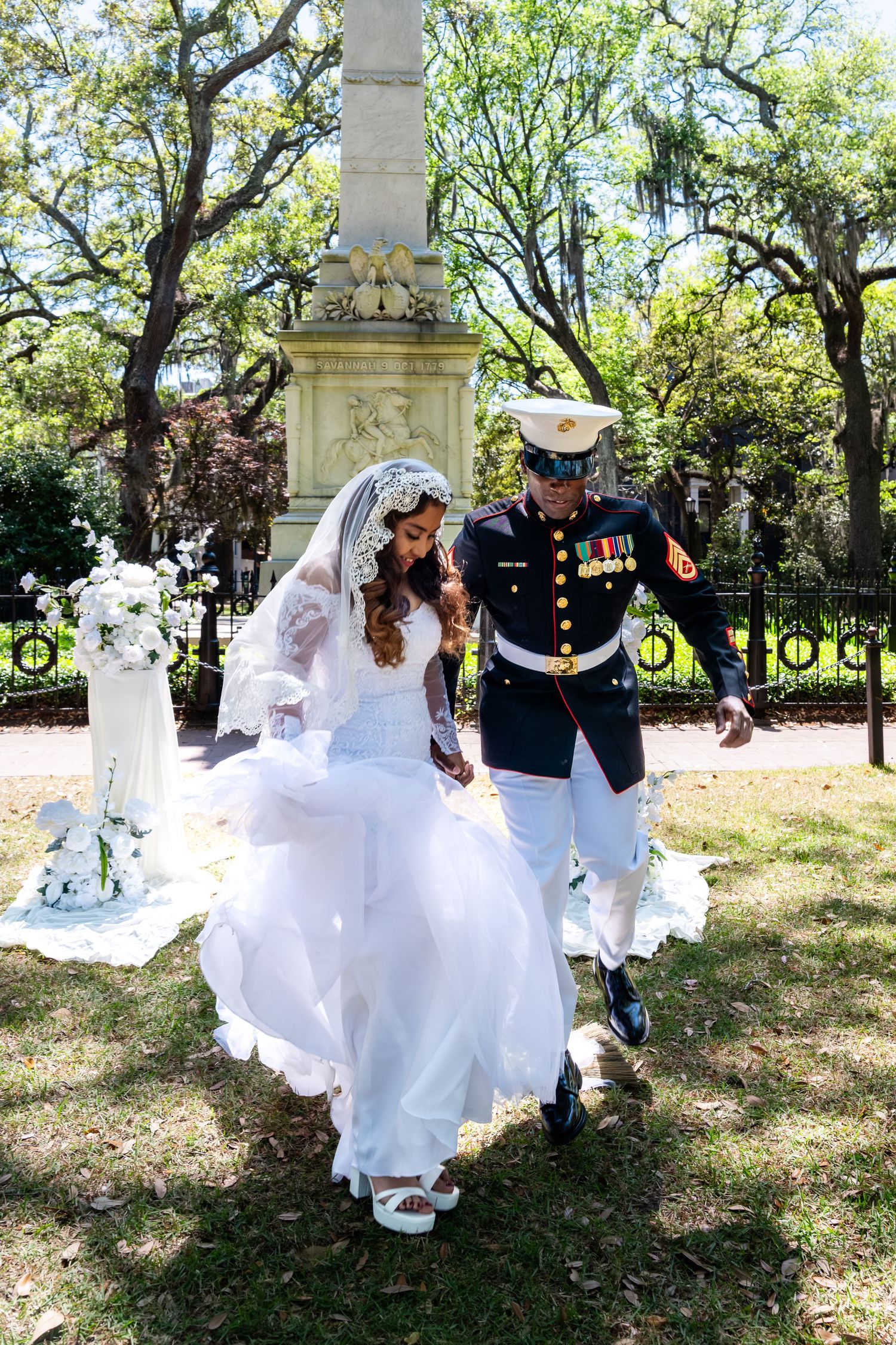 Marine groom in dress blues and bride in flowing white gown walk together through a sunlit park after ceremony.