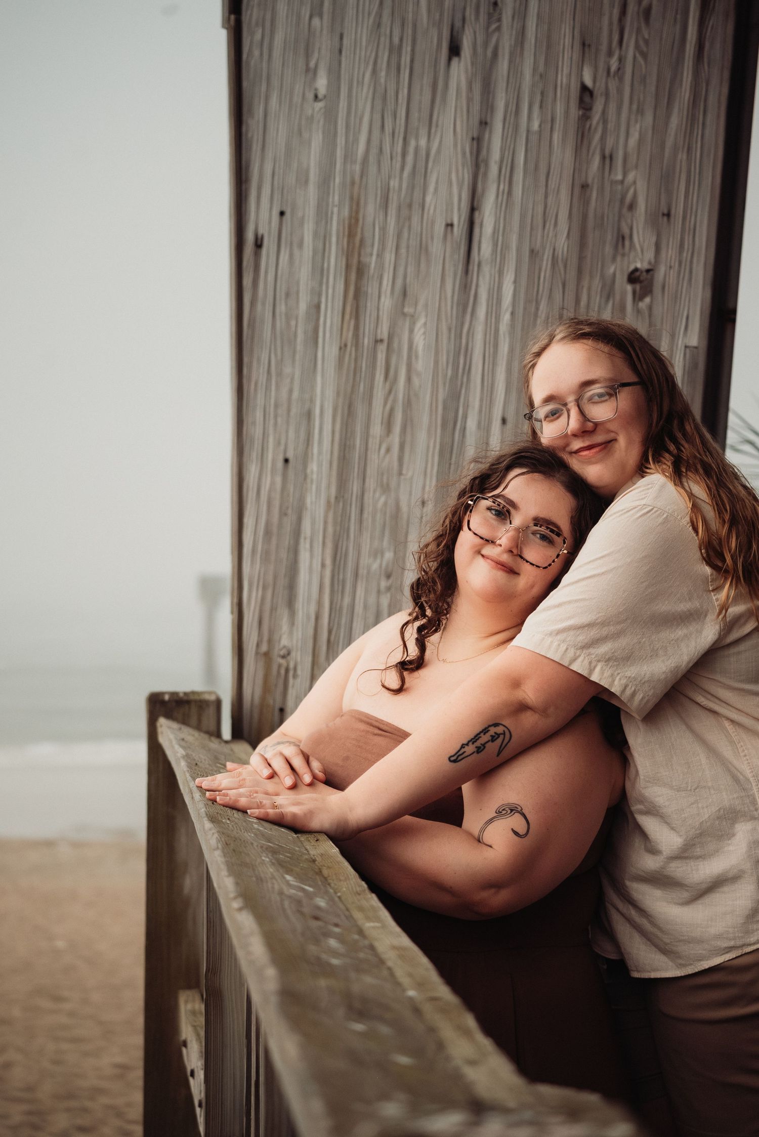 Two smiling women posing together leaning on a wooden railing with the beach visible in the background.