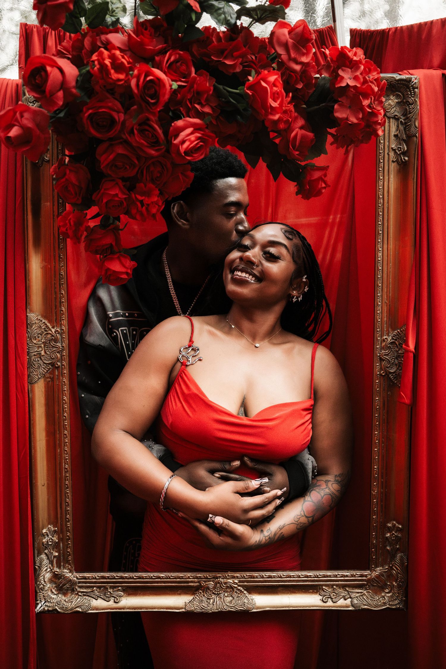 Couple posing in a golden frame surrounded by red roses and curtains, man kissing woman's cheek.