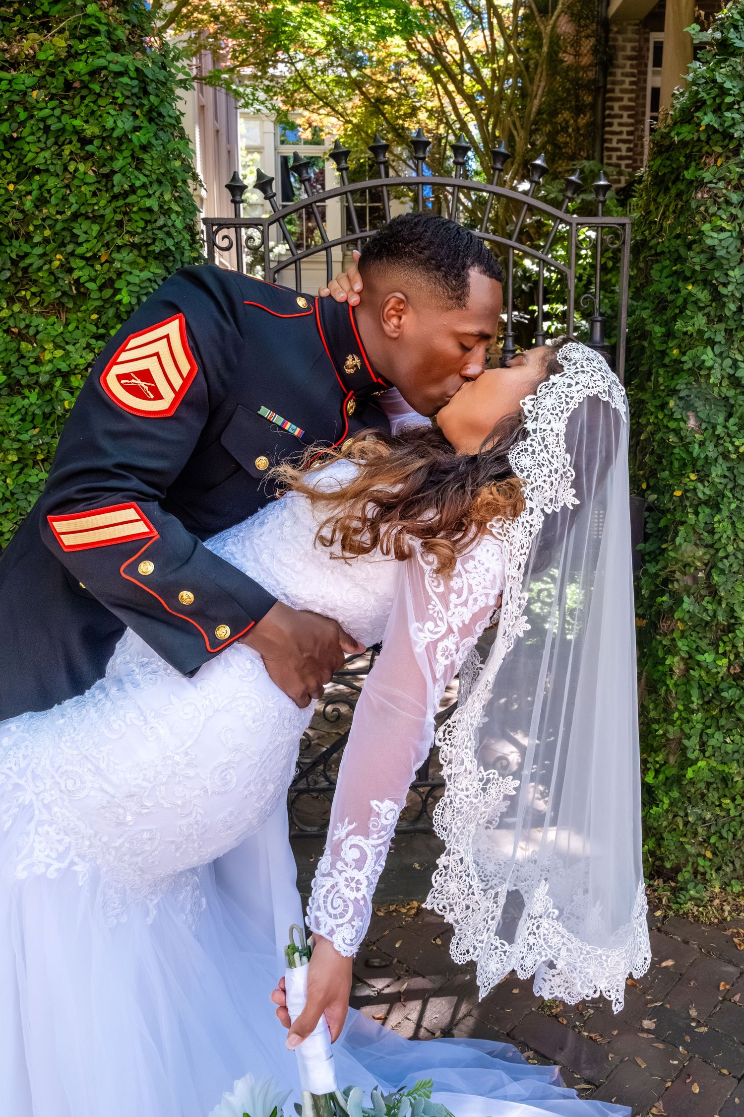 Marine groom in dress blues dips bride for romantic kiss at garden gate wedding photo.