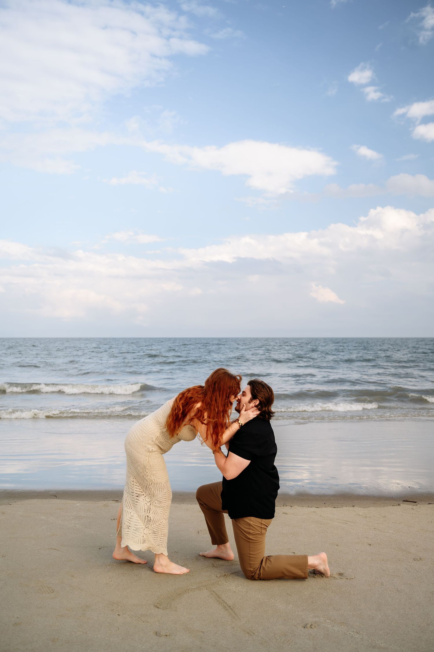 Man proposing on one knee to redhead woman in white dress on a sandy beach at sunset.