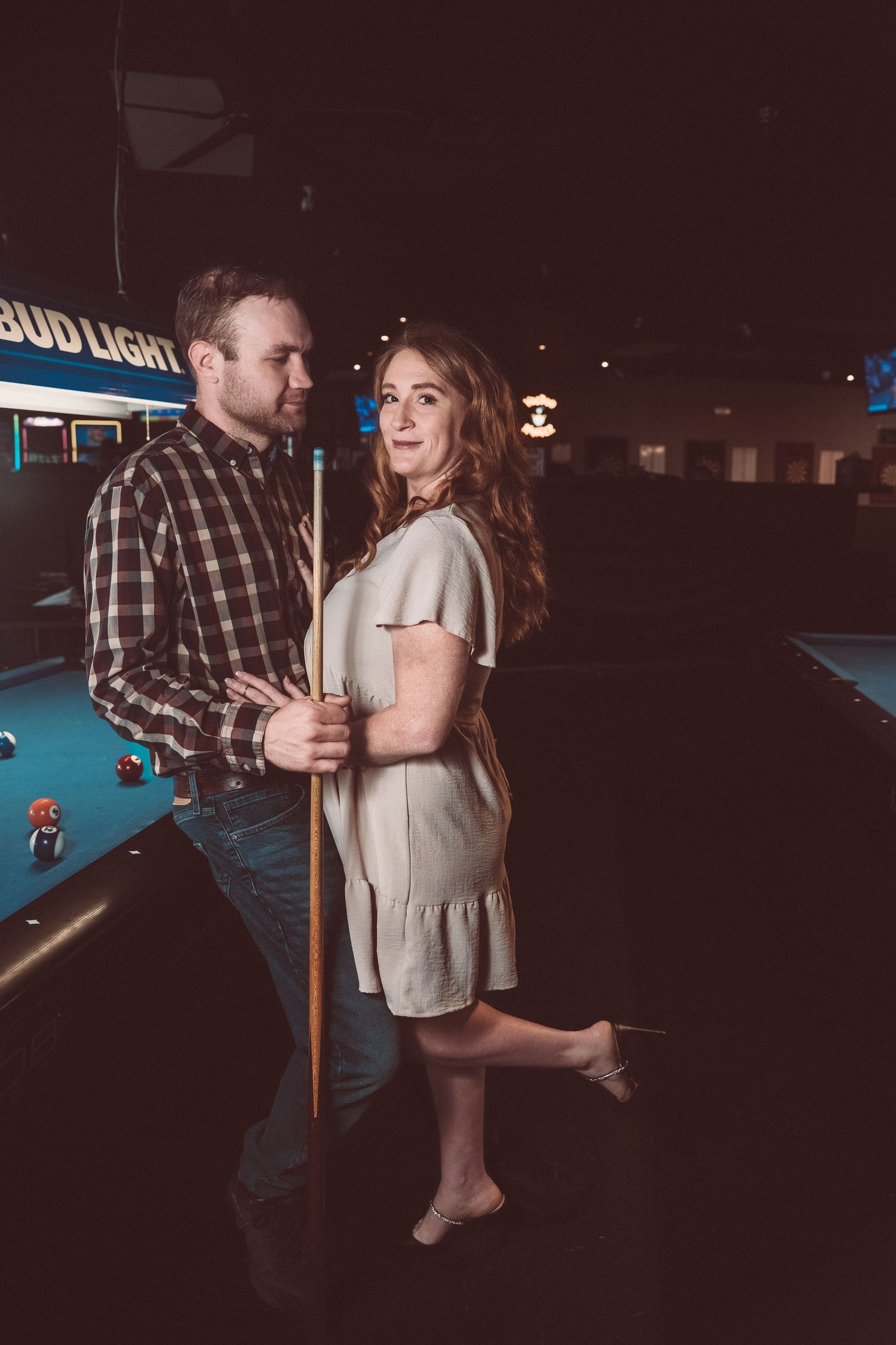 Couple sharing a romantic moment at a pool table in a dimly lit bar, holding a billiard cue together.