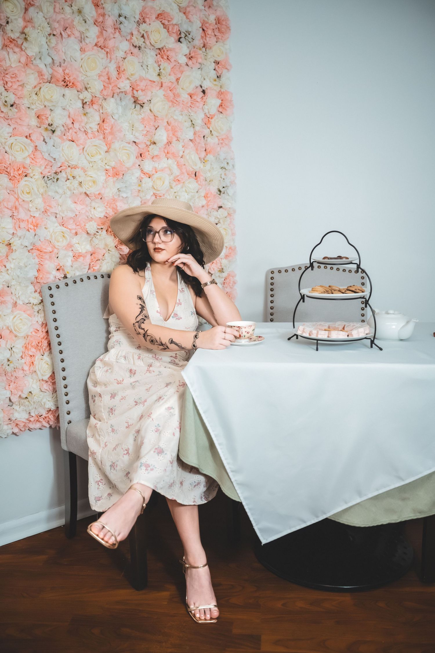 Woman in floral dress enjoys afternoon tea at table with pink flower wall backdrop and tiered serving tray.