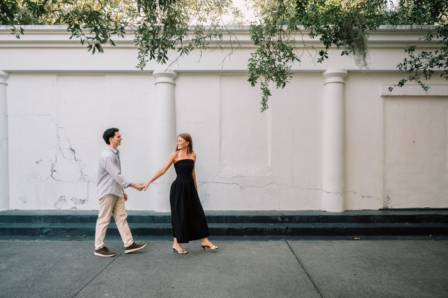 Engaged couple holding hands and walking along a white wall with hanging greenery above.