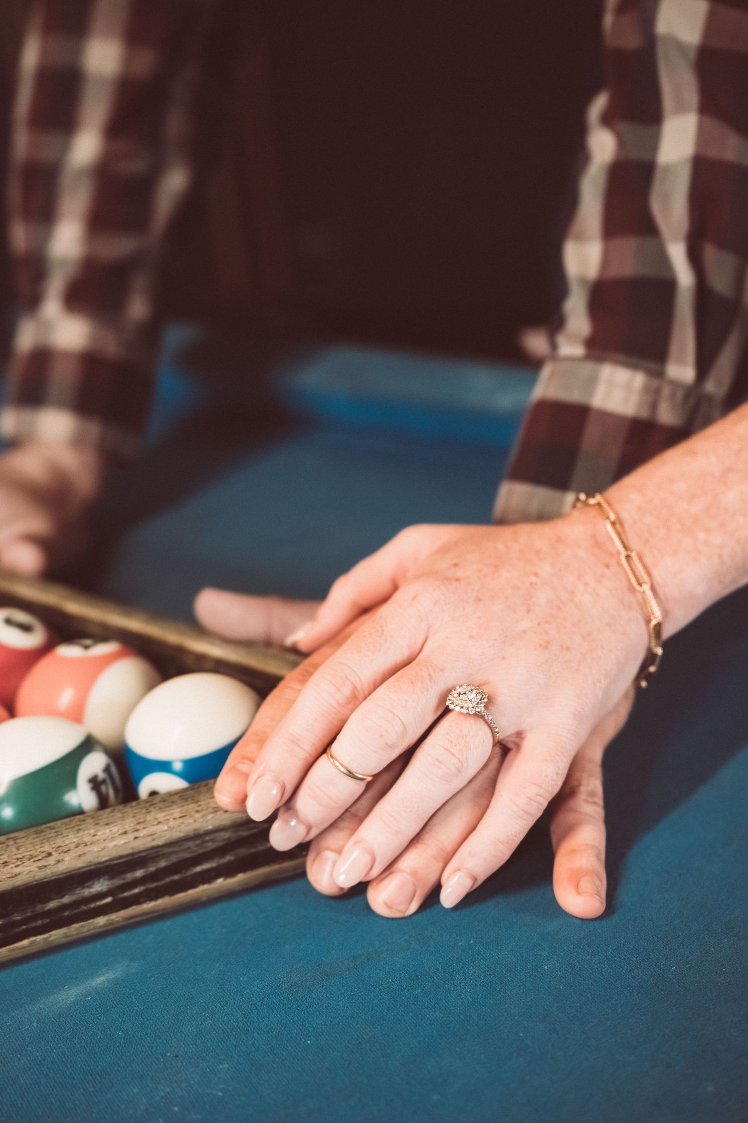 Close-up of a womans hand wearing a diamond engagement ring resting on a pool table near billiard balls.
