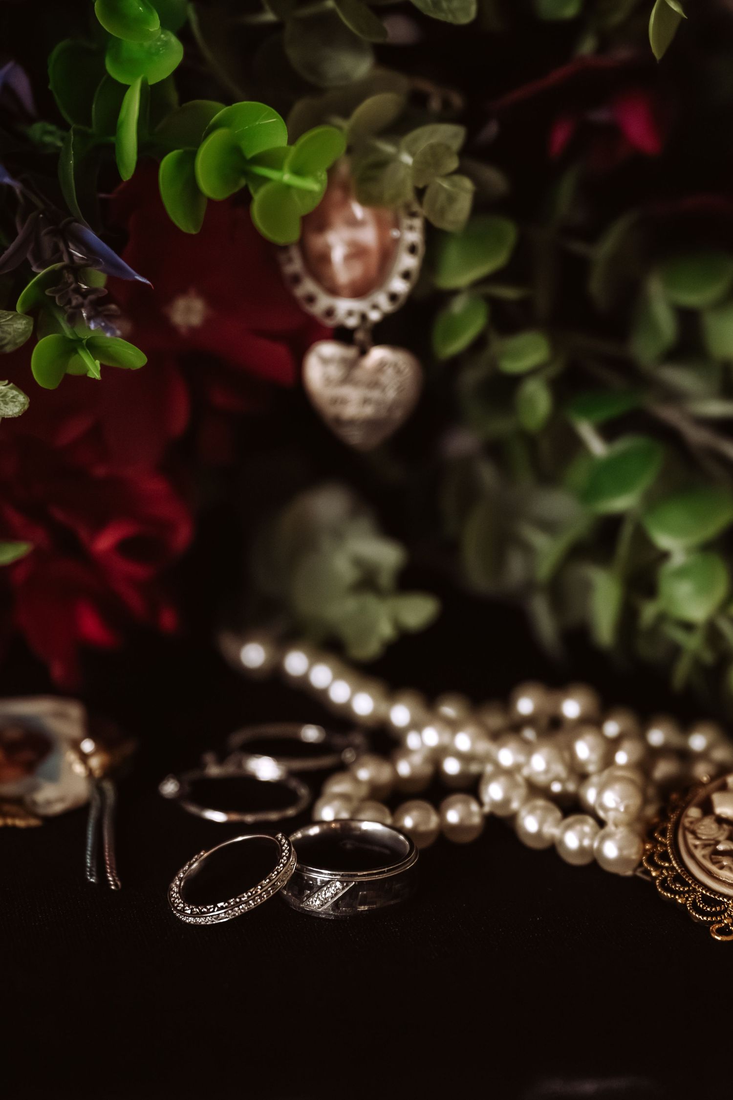 Wedding rings and pearl necklace displayed beside a bouquet of red roses and greenery on a dark surface.