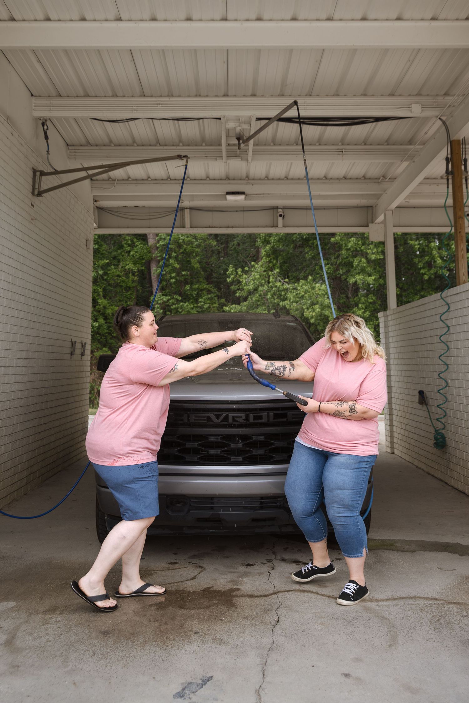 Two women in pink shirts playfully pulling a hose at a self-service car wash.