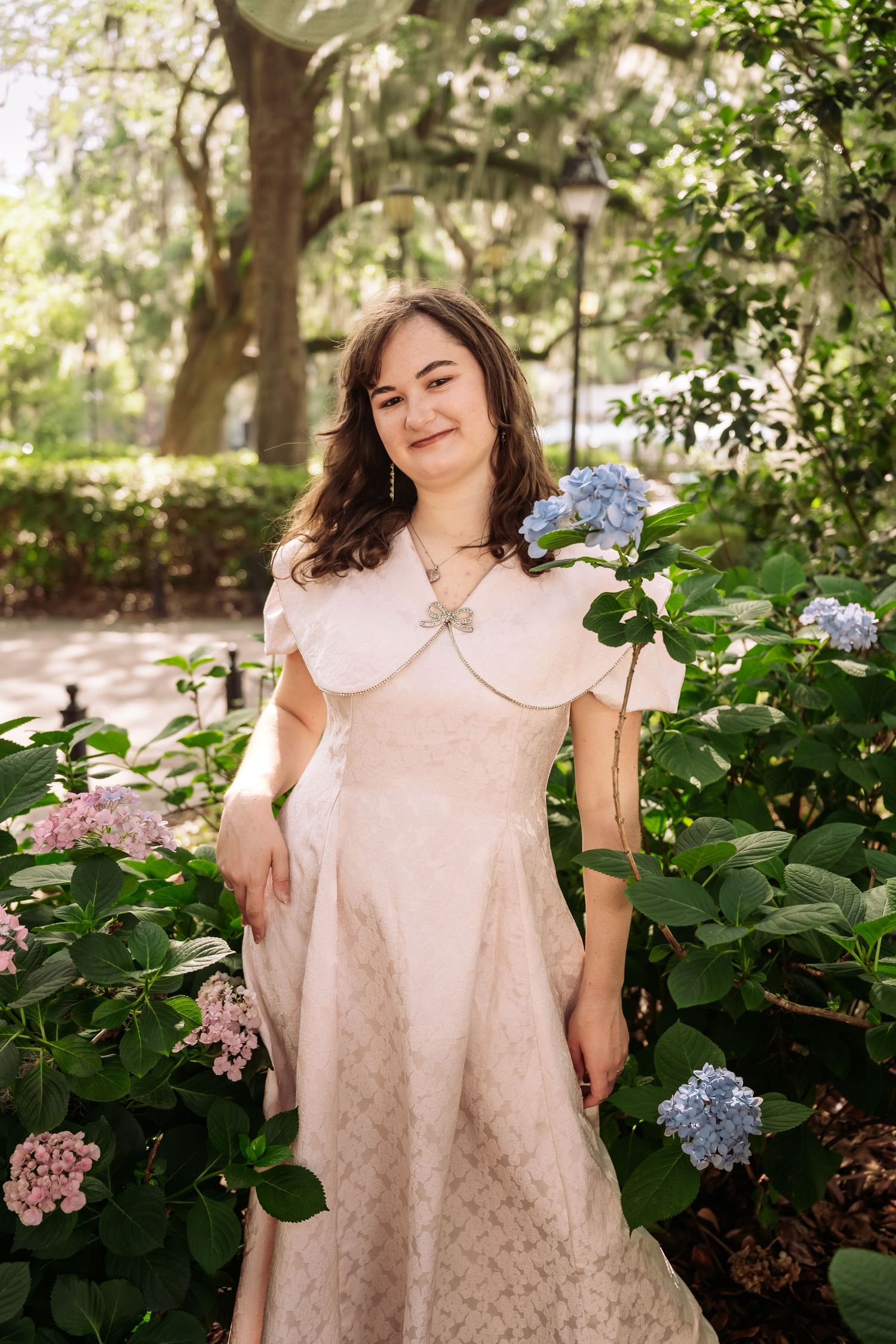 Young woman in vintage pink dress posing among blooming hydrangeas in a sunny park.