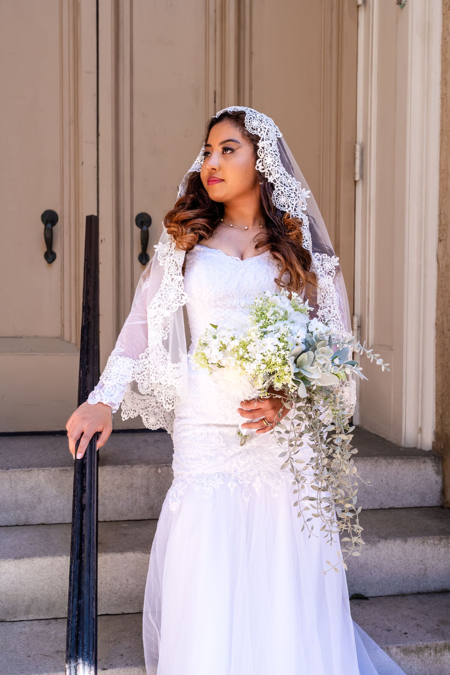 Elegant bride in lace mermaid gown and mantilla veil holds white floral bouquet on church steps.
